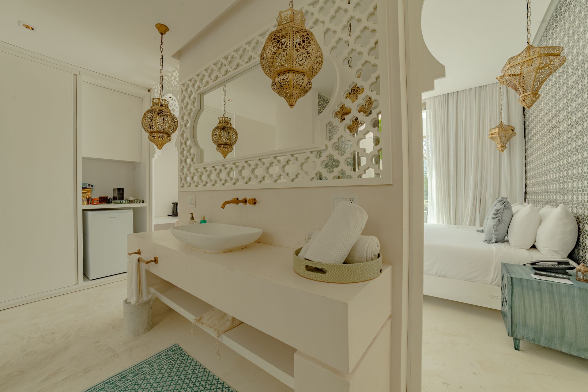 Baño y dormitorio en blanco y dorado con lámparas colgantes ornamentadas y una mesa auxiliar color turquesa.