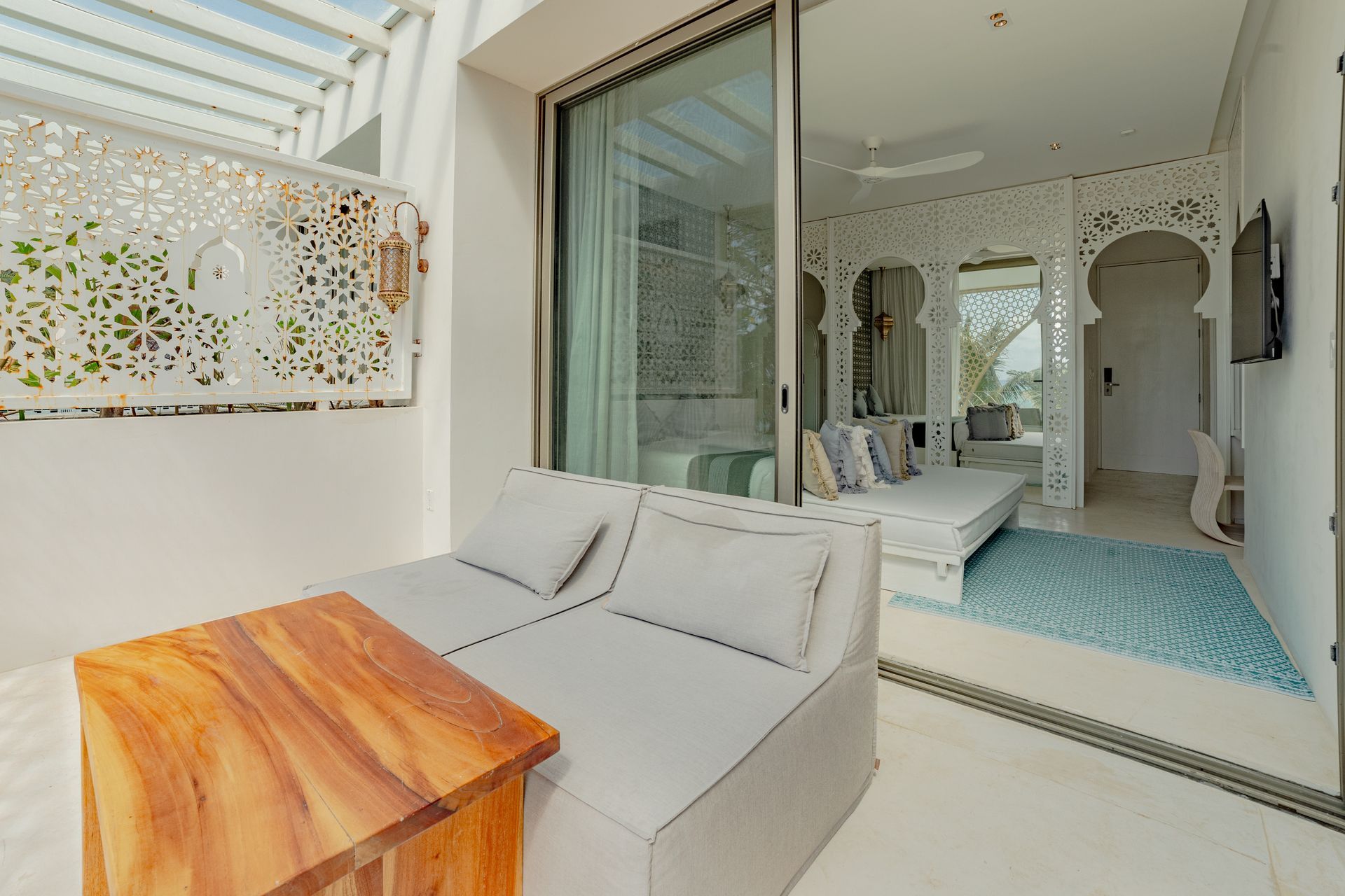 Zona de estar al aire libre con sofá blanco, mesa de madera y puertas corredizas de vidrio que dan a un dormitorio.