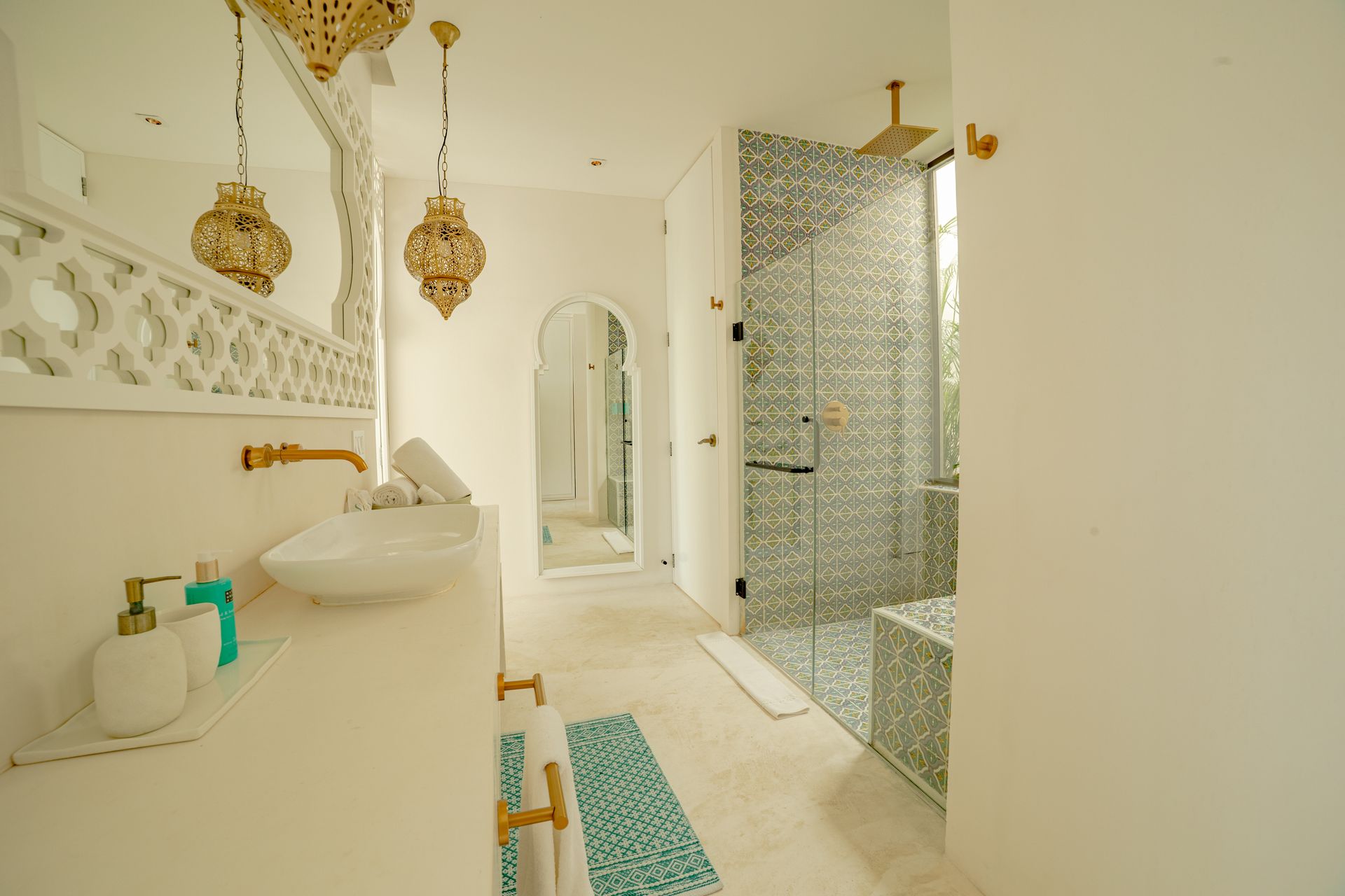 Baño blanco con espejos ornamentados, accesorios dorados y ducha con azulejos de mosaico.