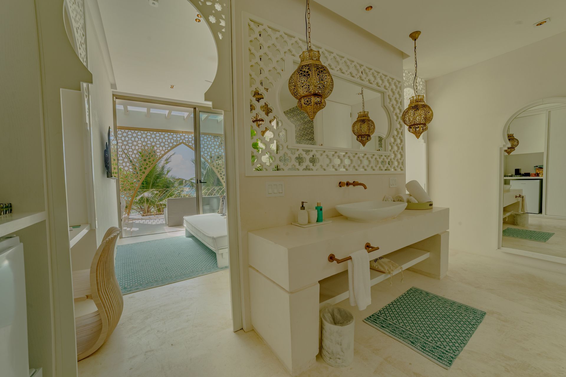 Baño con paredes blancas, espejo ornamentado, lámparas doradas y puerta abierta a un dormitorio con vista al mar.