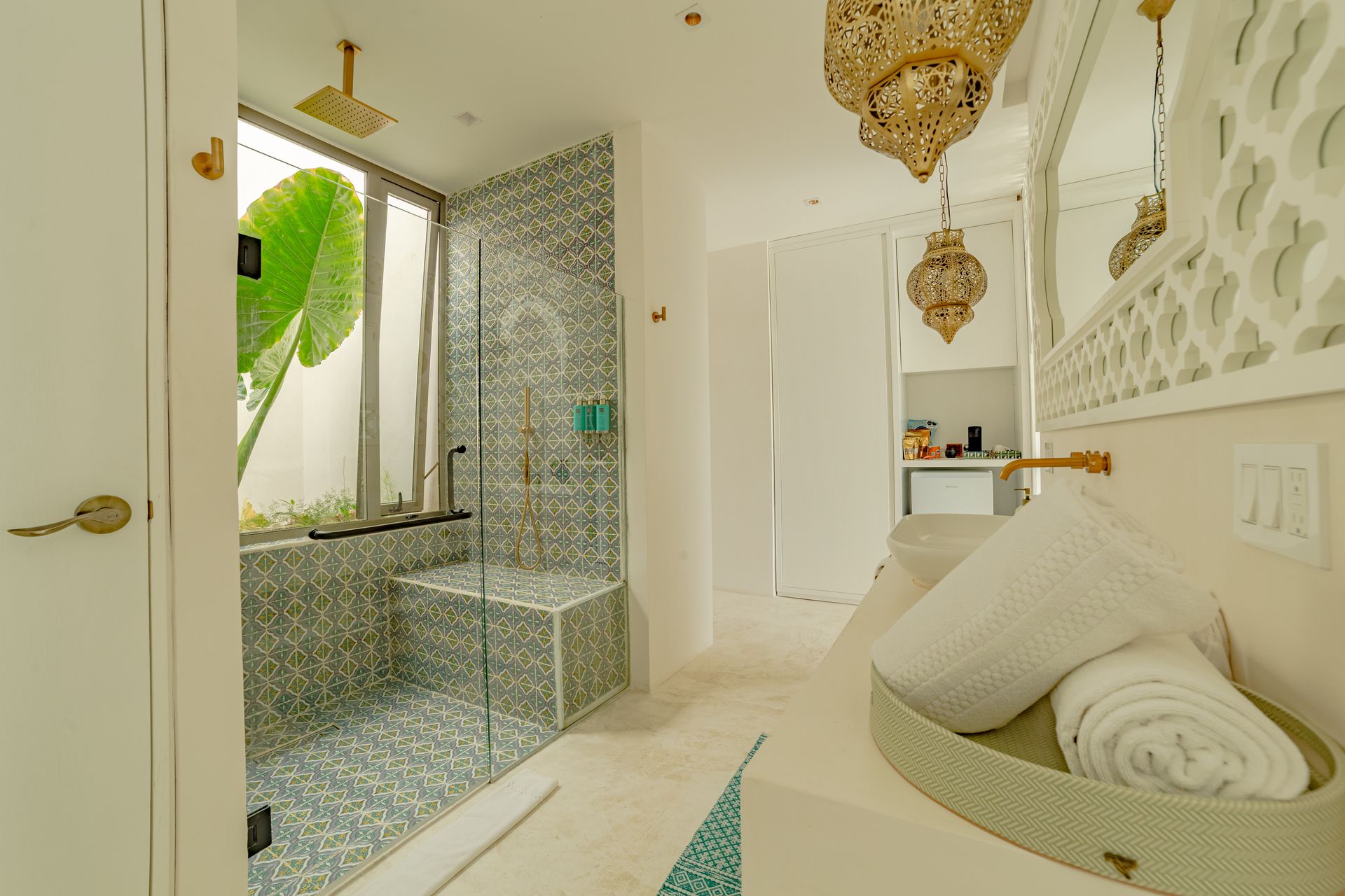 Baño de estilo marroquí. Ducha con azulejos de mosaico, banco, lámparas ornamentadas y lavabo con toallas.
