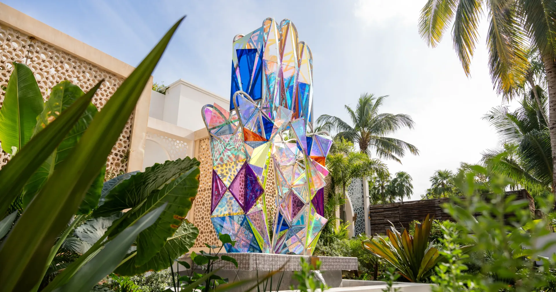 Escultura de vidriera colorida de una mano abierta en un entorno de jardín, con follaje verde y palmeras.