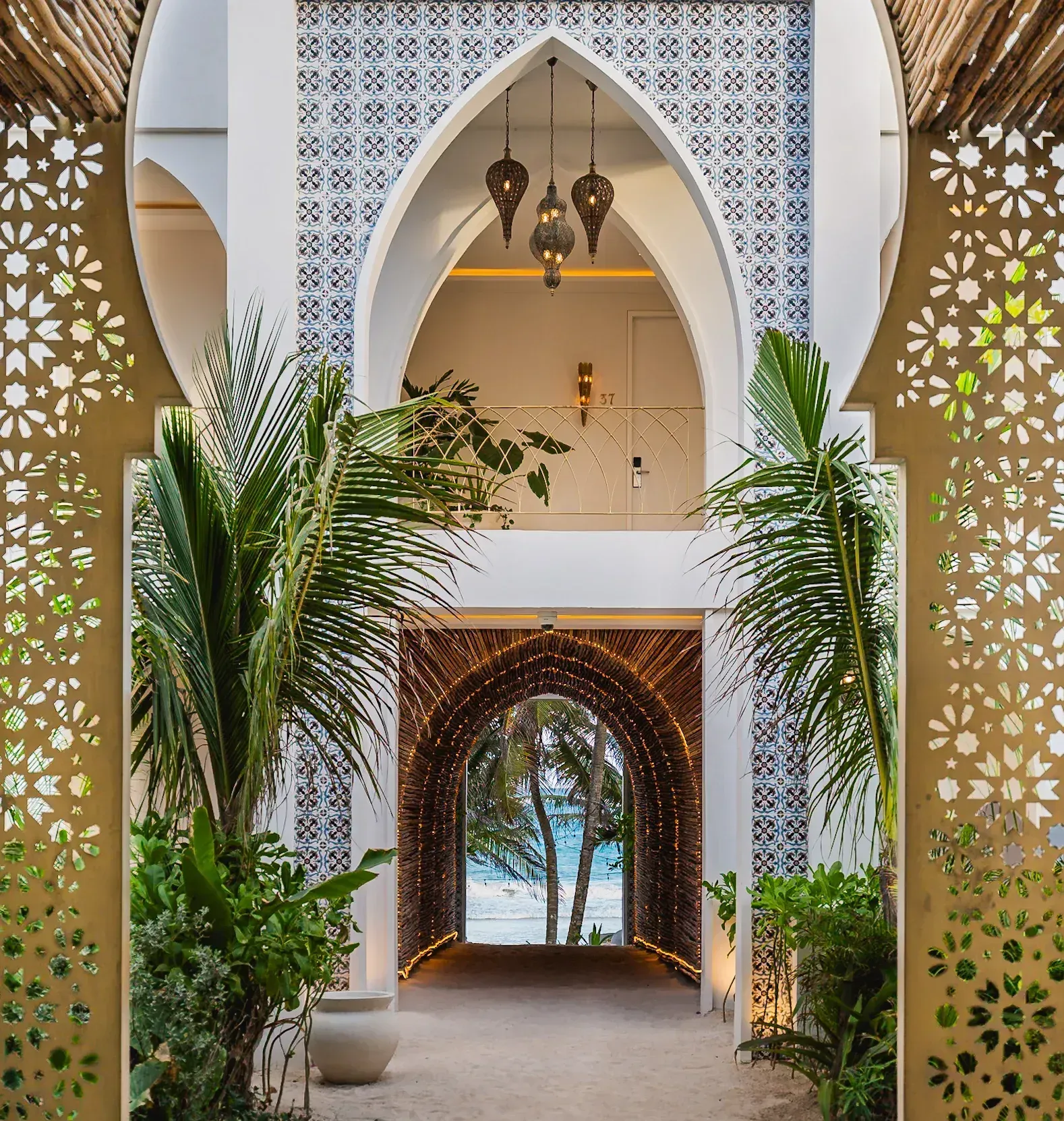 Puerta arqueada adornada que conduce a una vista de la playa con follaje tropical y paredes blancas estampadas.