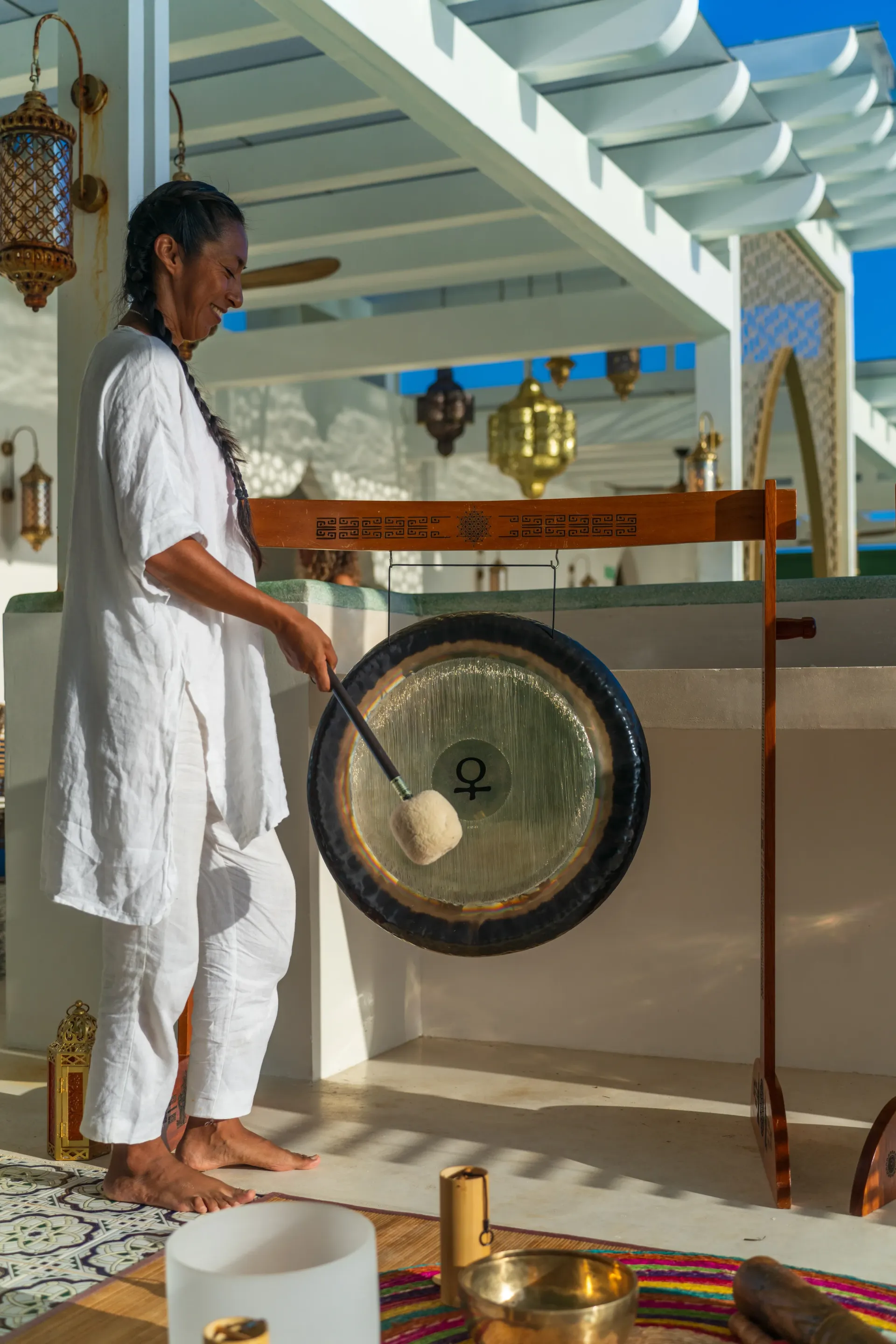 Woman in white clothes striking a gong with a mallet outdoors.