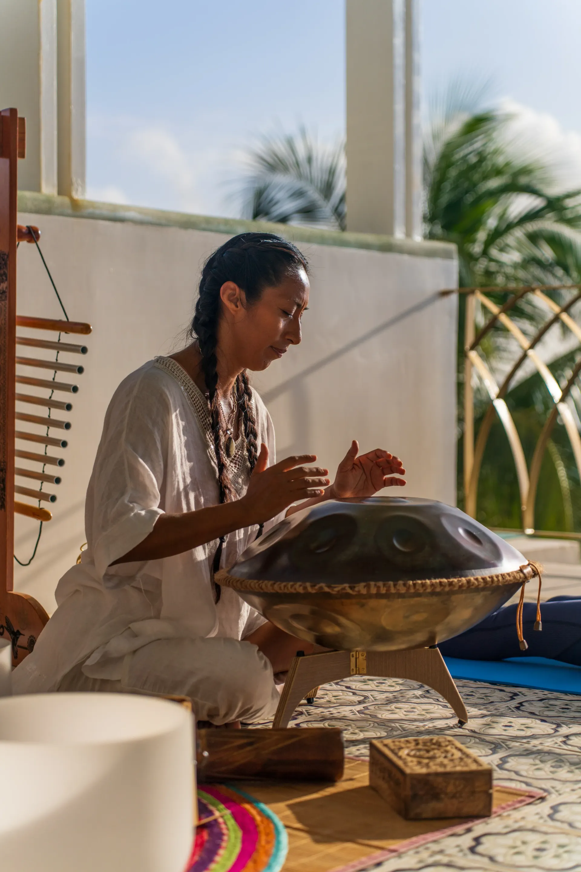 Una mujer toca un tambor handpan al aire libre. Está sentada, vestida de blanco, concentrada. Se ven la luz del sol y la vegetación.