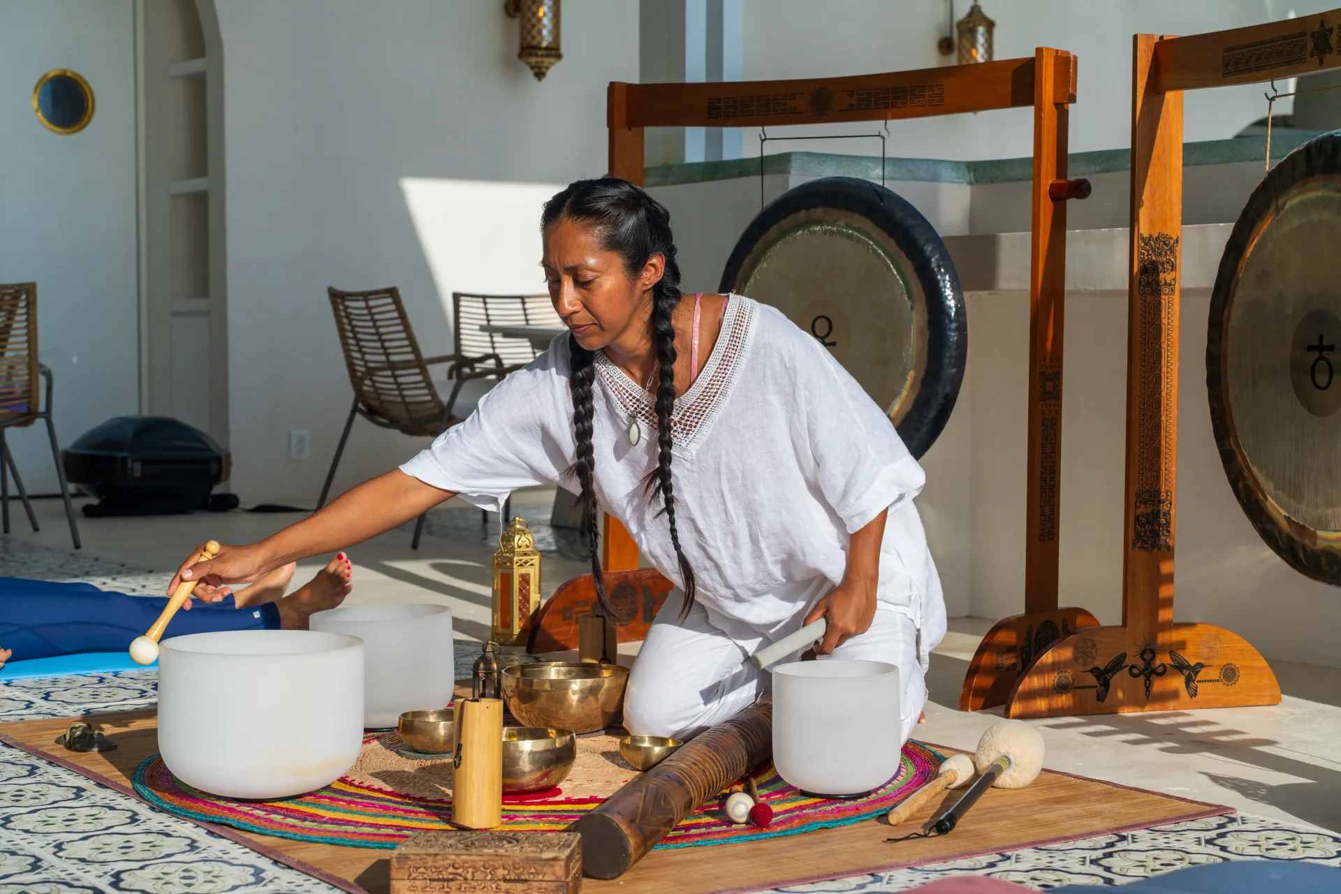 Woman plays crystal bowls and gongs in a white-walled space with meditation tools.
