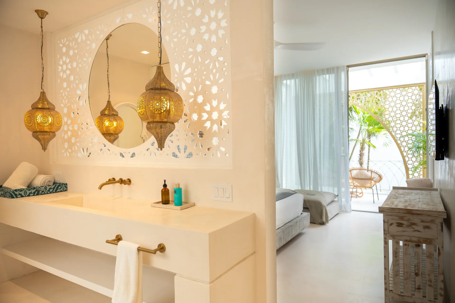 Baño con luces colgantes doradas ornamentadas, pared blanca tallada y vista al dormitorio.