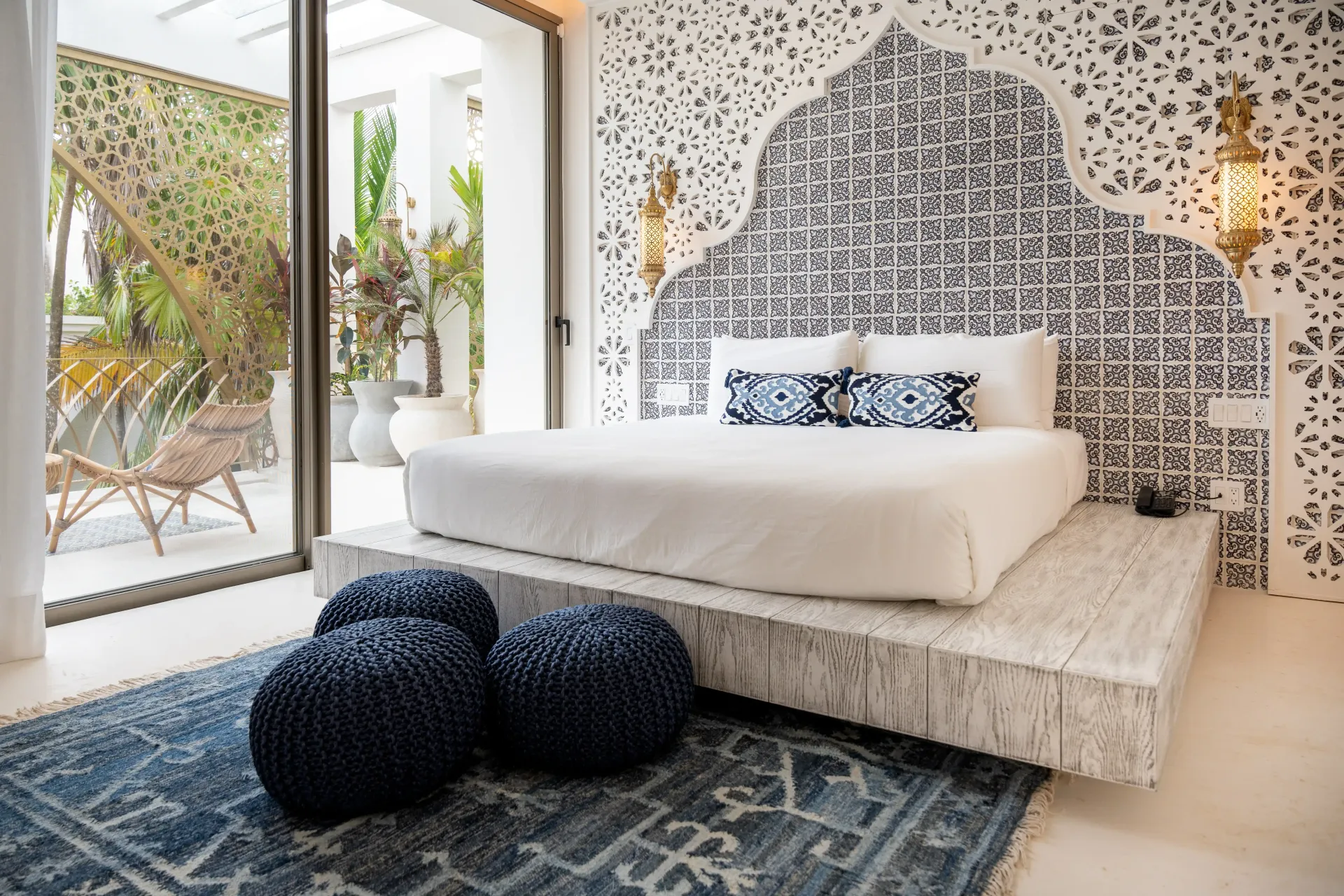 Dormitorio con cabecera ornamentada, ropa de cama blanca, alfombra azul y puerta abierta a un patio exterior.
