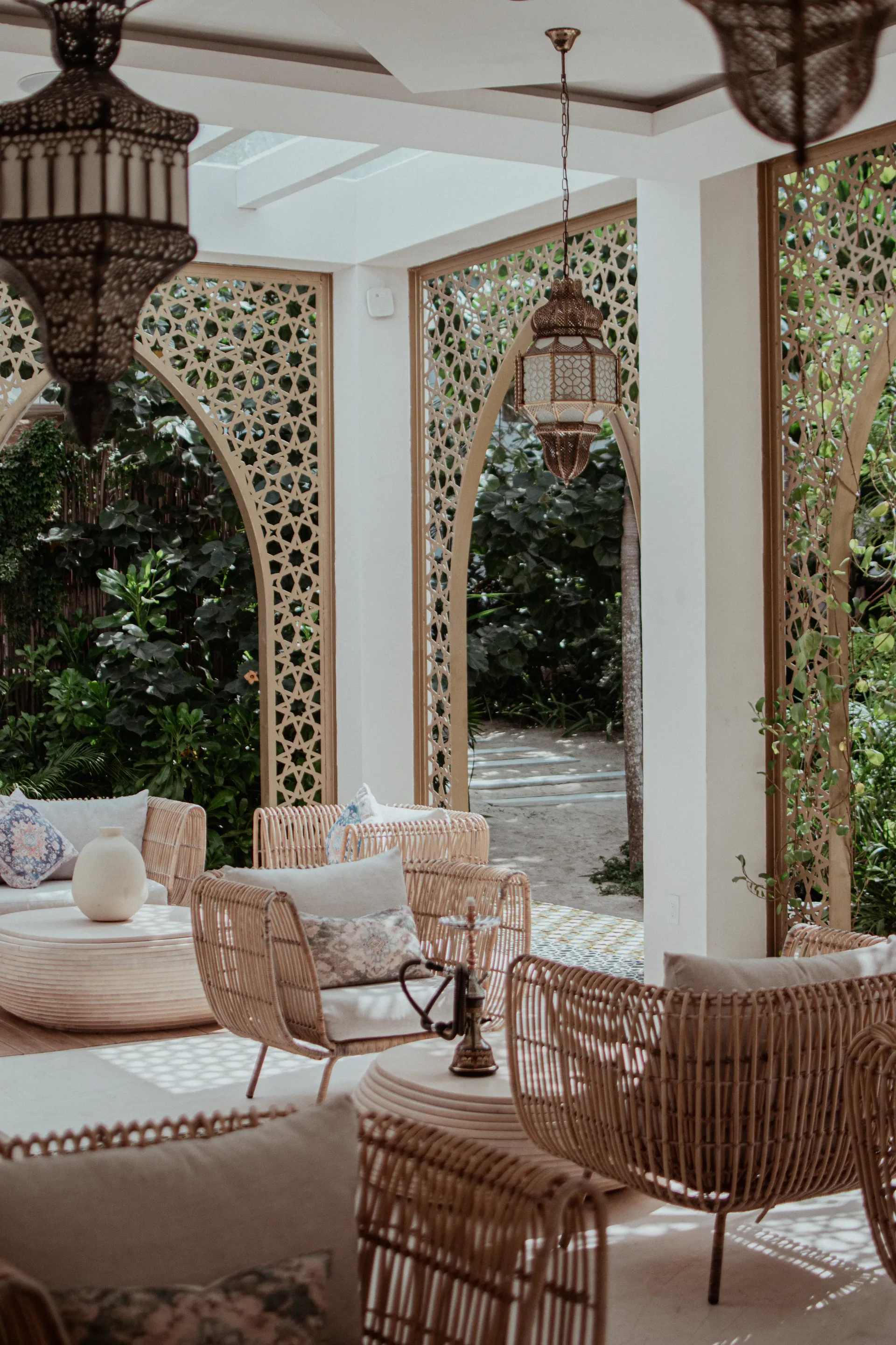 A sunlit outdoor lounge with wicker chairs, intricate arched lattice screens, and hanging Moroccan-style lanterns.