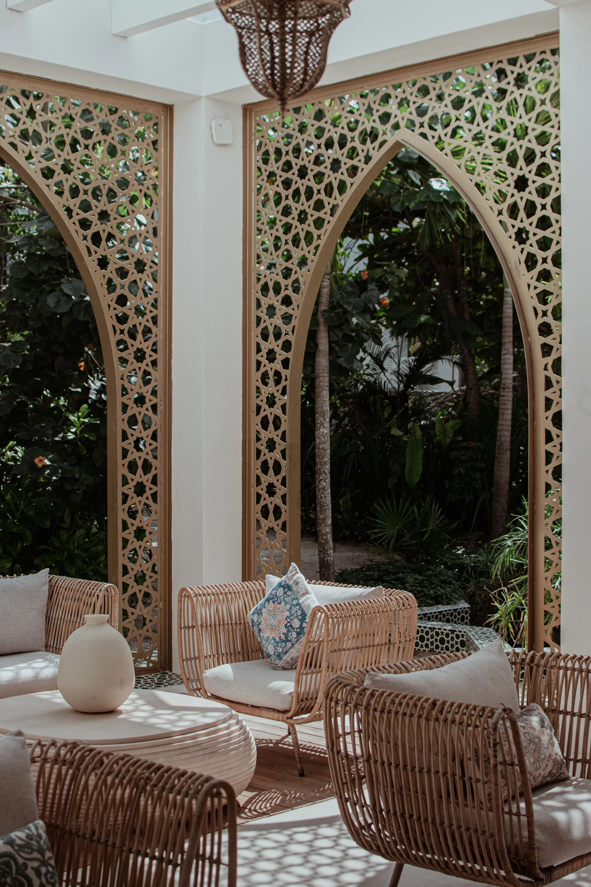 A sunlit patio featuring wicker furniture, geometric arched screens, and lush greenery in a serene, neutral-toned space.
