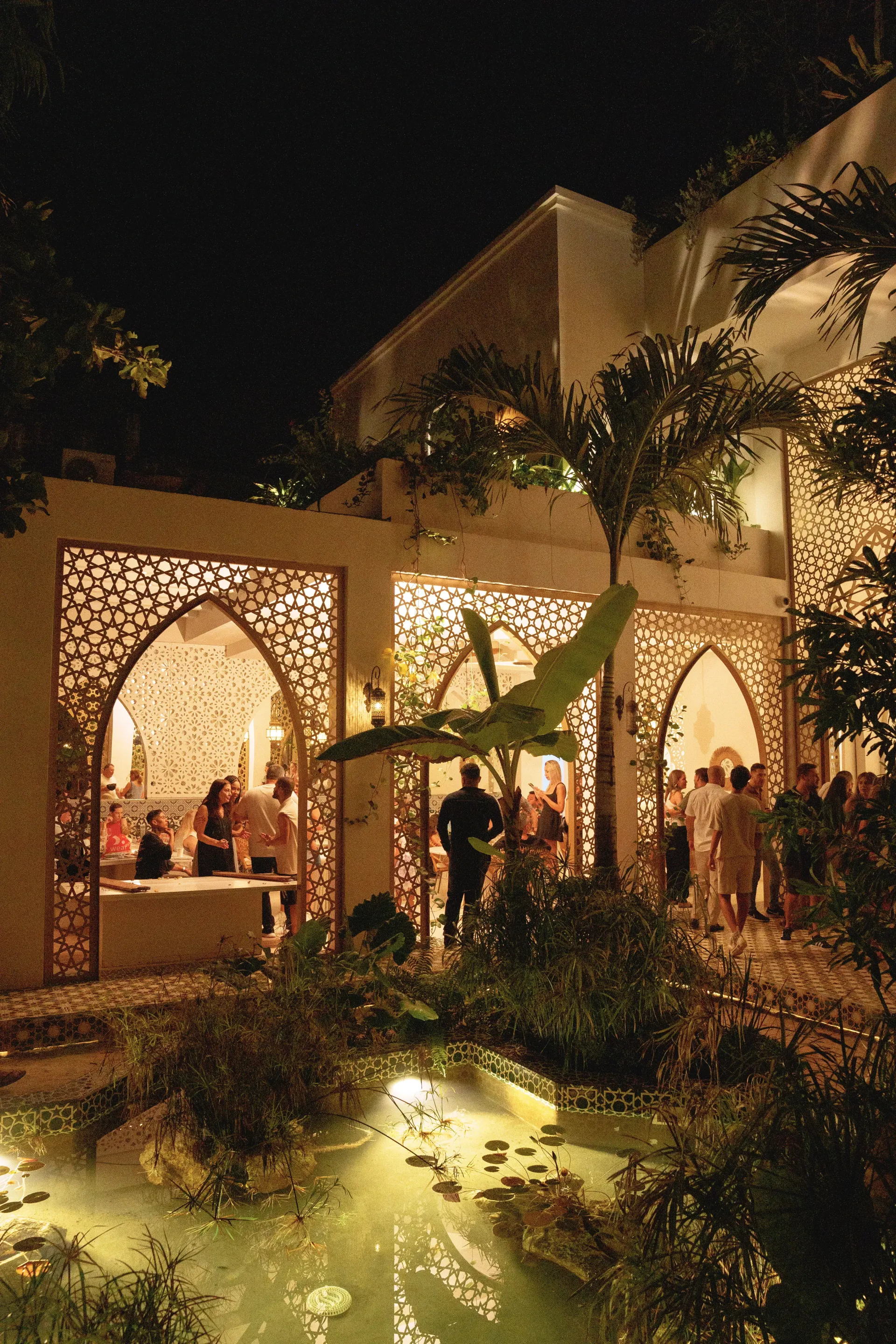 A night scene showing a building with ornate, arched windows illuminated from within, overlooking a small, lit garden pool.
