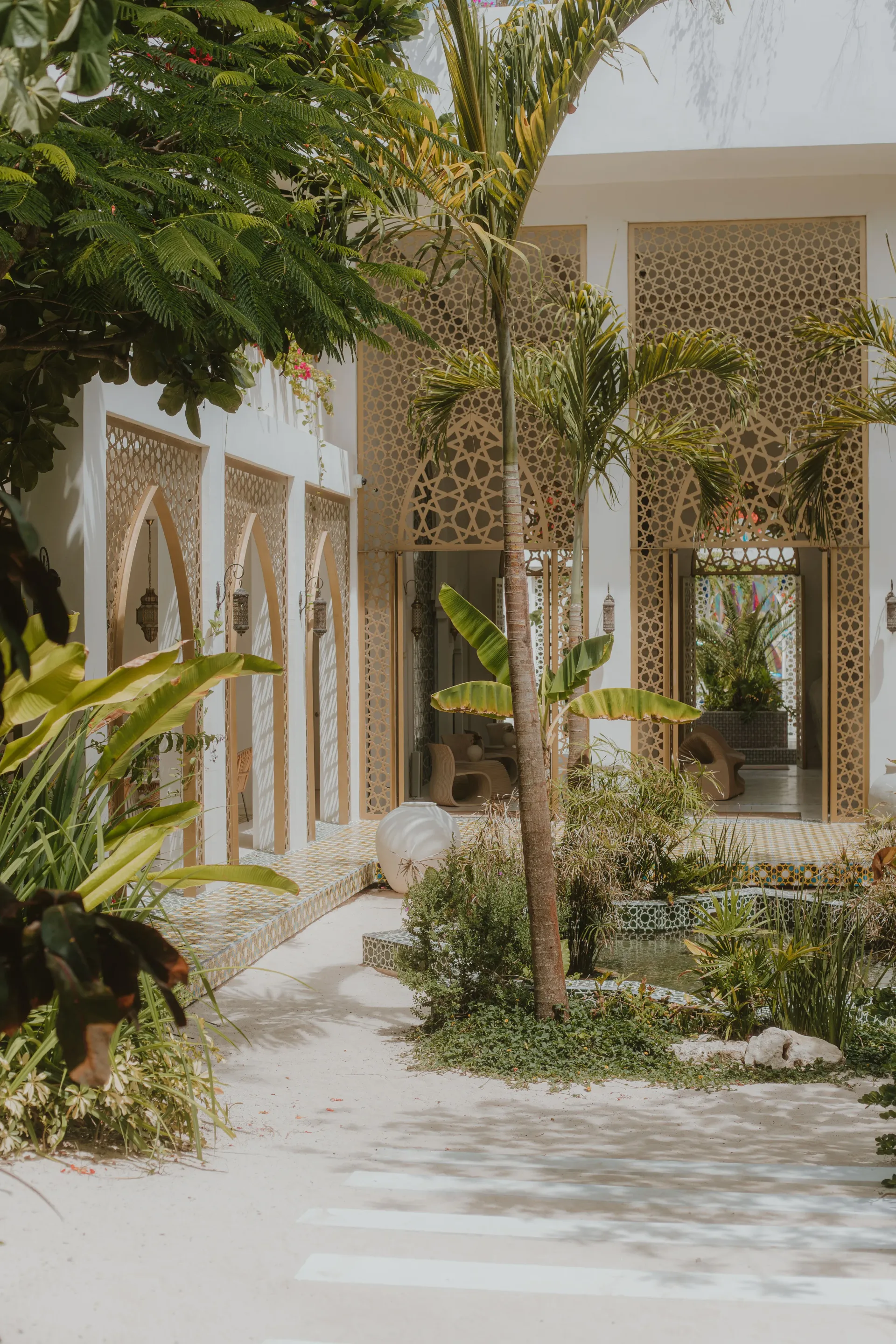 A sunlit tropical courtyard featuring white arched walls with intricate lattice patterns, lush green plants, and a palm.