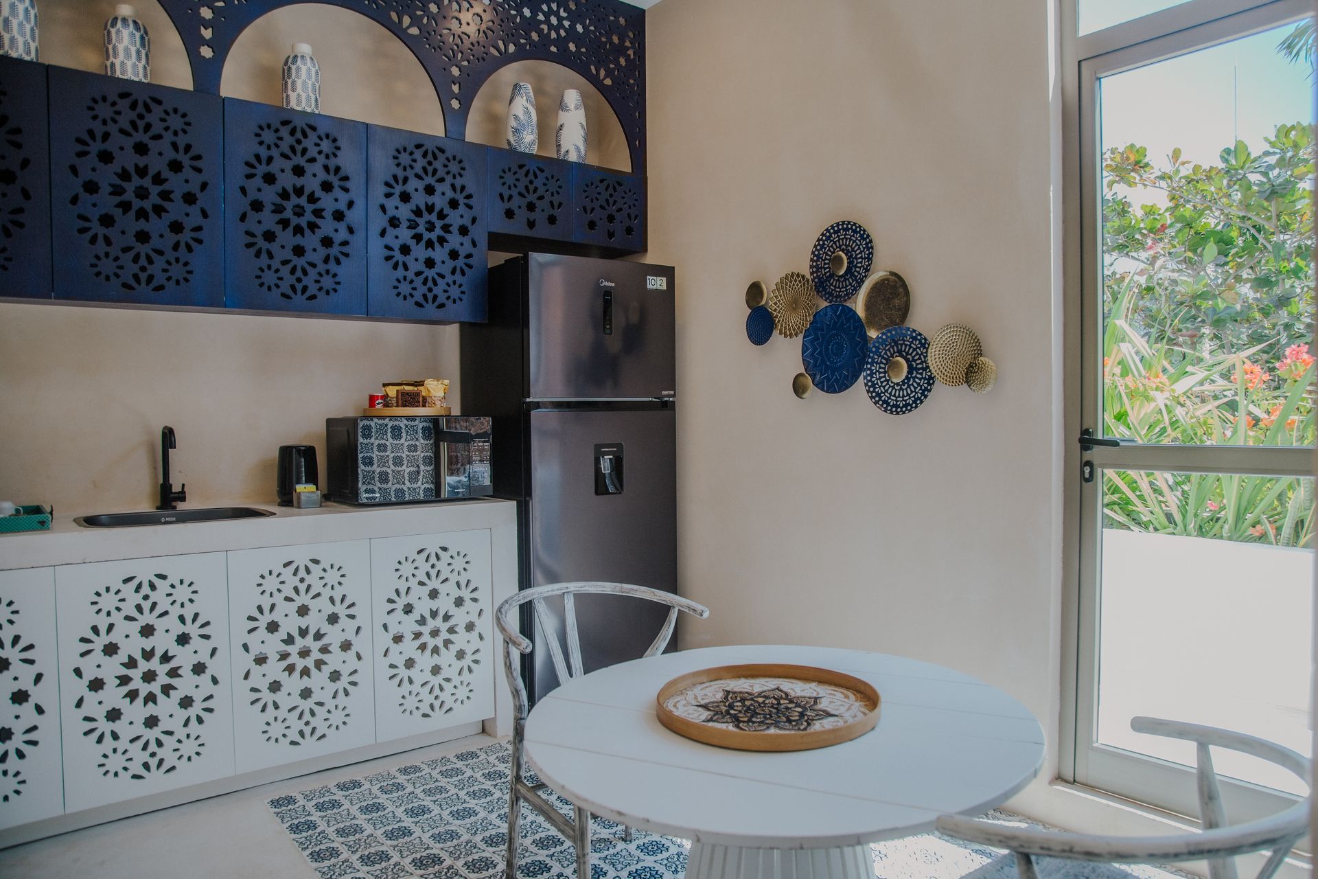Una cocina con armarios blancos con elaborados detalles calados, estanterías de pared azul oscuro, una mesa redonda y una puerta de cristal que da al patio.