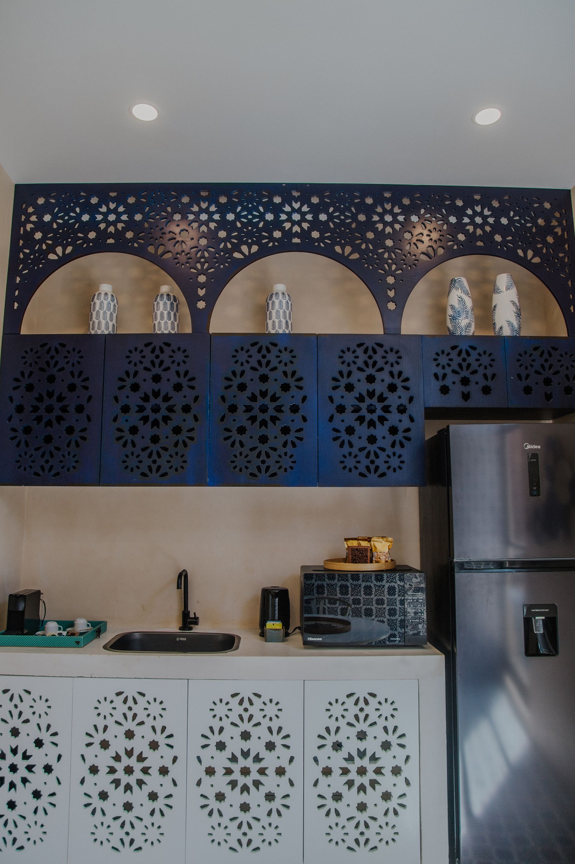 Una pequeña cocina con armarios superiores de color azul oscuro con intrincados detalles calados, estantes arqueados abiertos y base blanca.