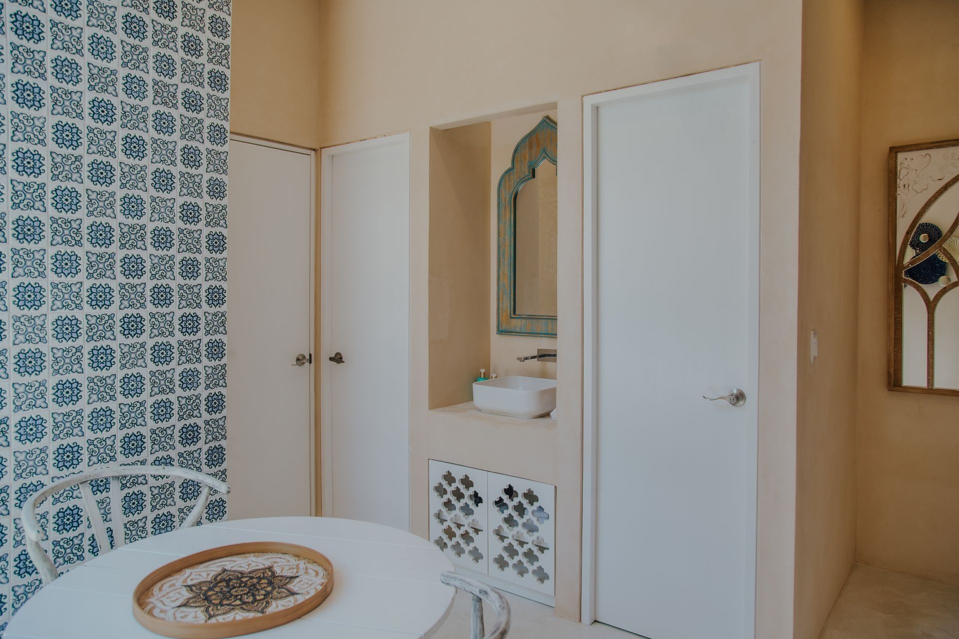 Una mesa circular con una bandeja decorativa se sitúa frente a una pared beige con un detalle de azulejos azules y blancos, un lavabo y una puerta.
