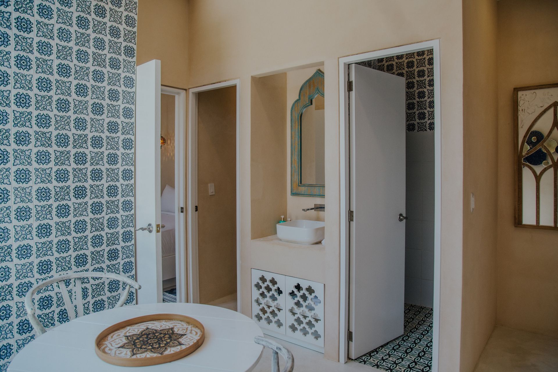 A white table with a woven tray sits before a blue-patterned wall, opening to a neutral-toned room with a vanity area.