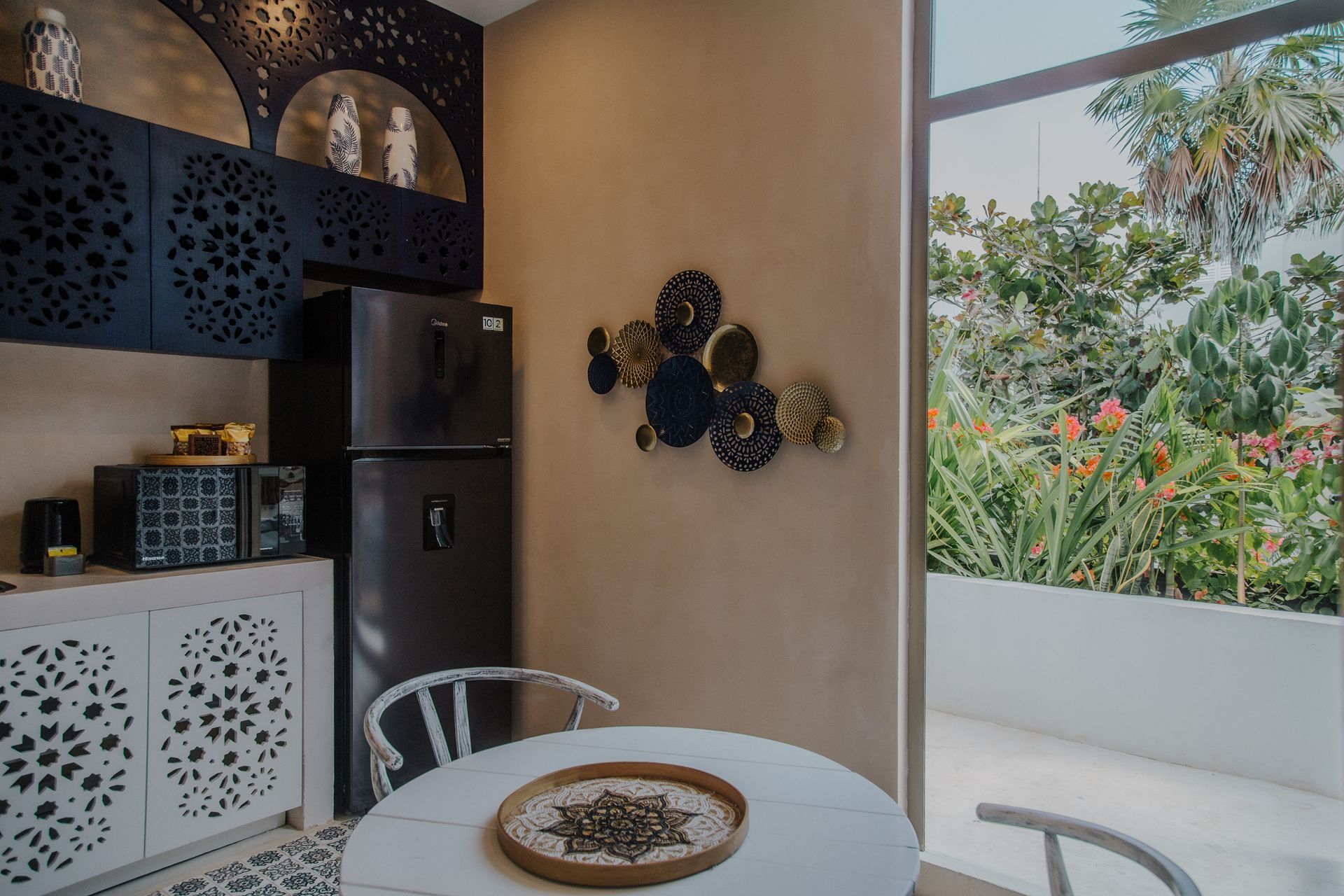 A cozy dining nook with a white table, patterned cabinets, a dark refrigerator, and a wall of lush tropical greenery.