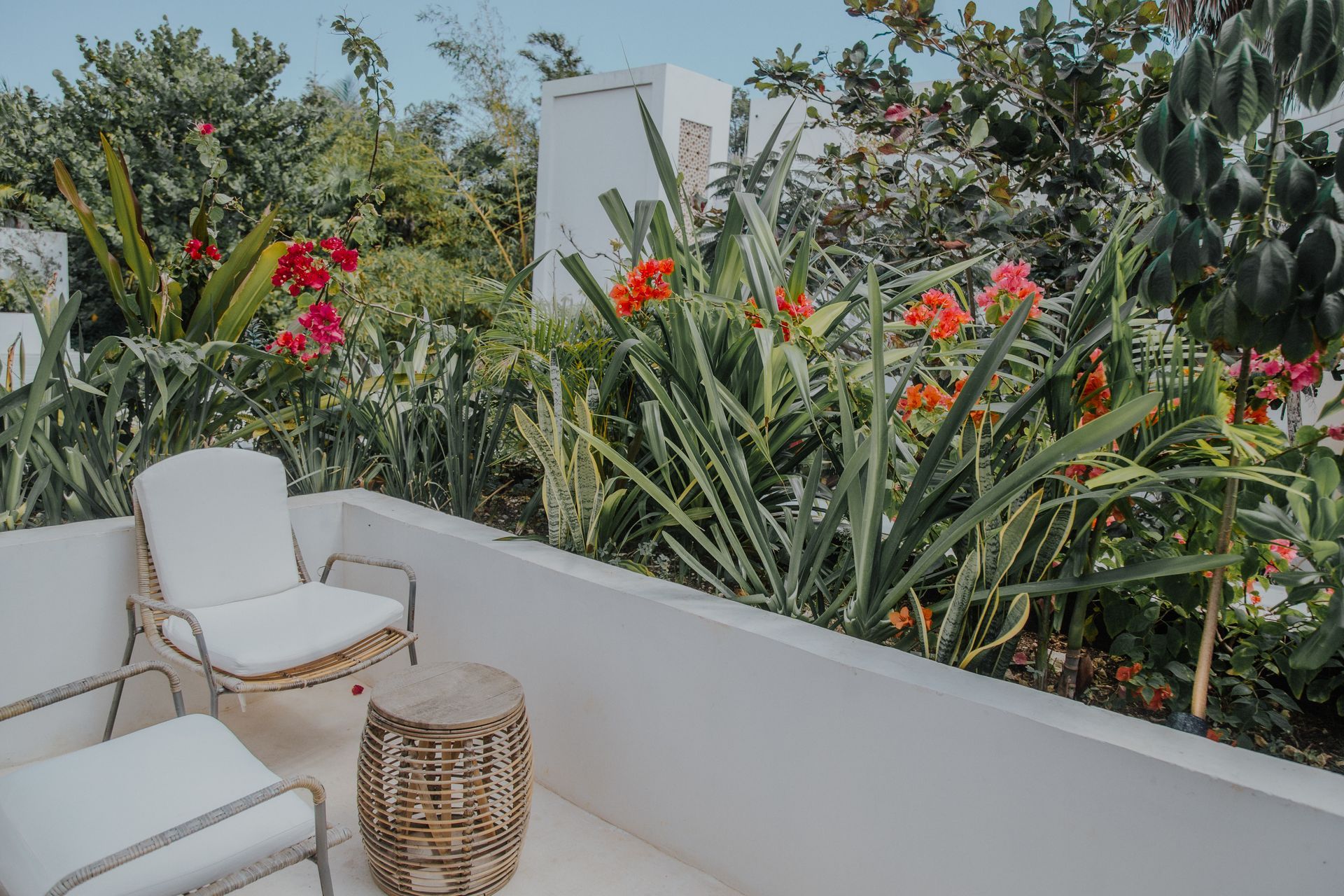 Dos sillas blancas modernas y una mesa auxiliar de mimbre en un balcón rodeado de exuberante vegetación y flores de color naranja brillante.