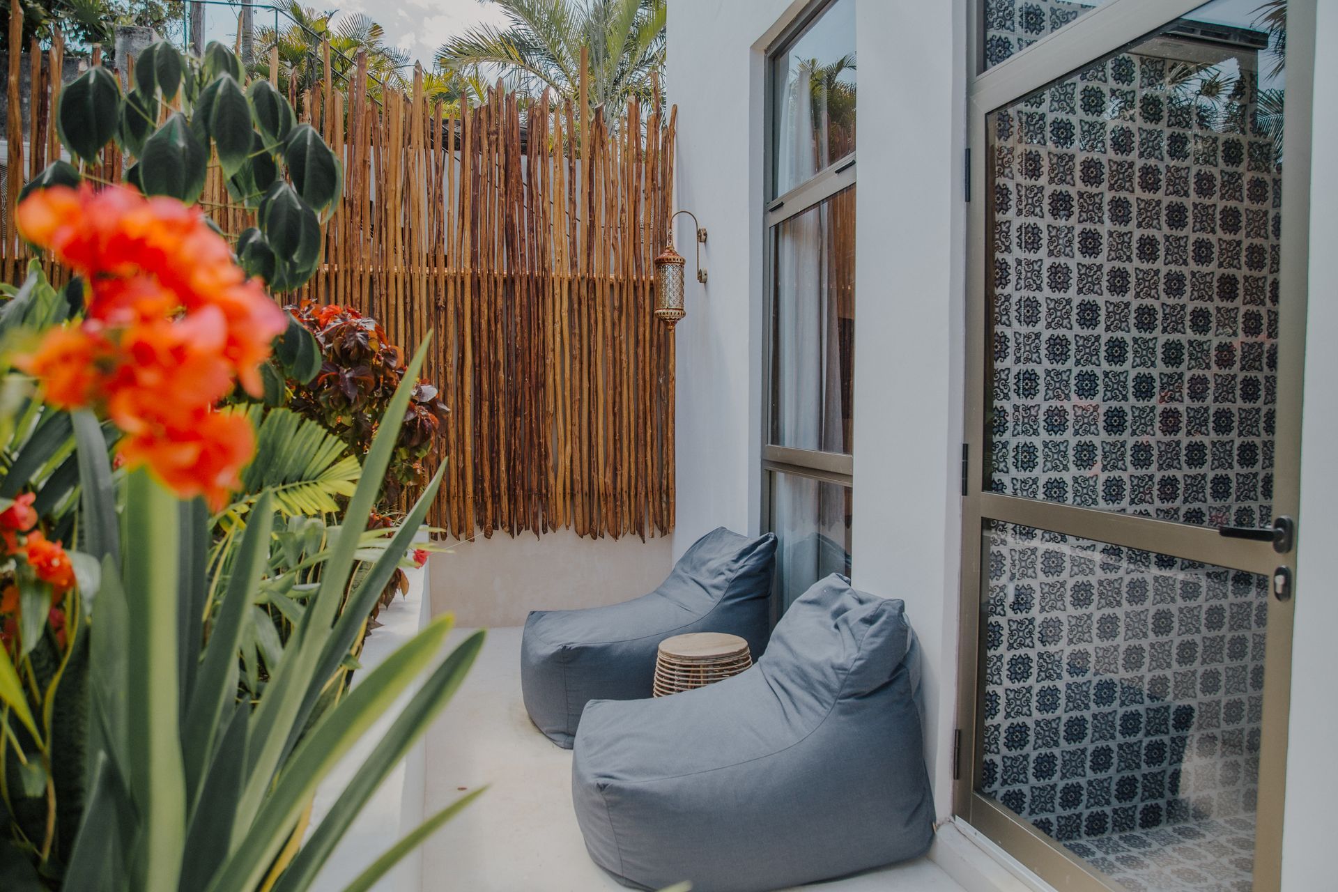 Dos sillones tipo puf grises se encuentran en un patio junto a una puerta de cristal, con flores de color naranja vibrante y una valla de listones de madera cerca.