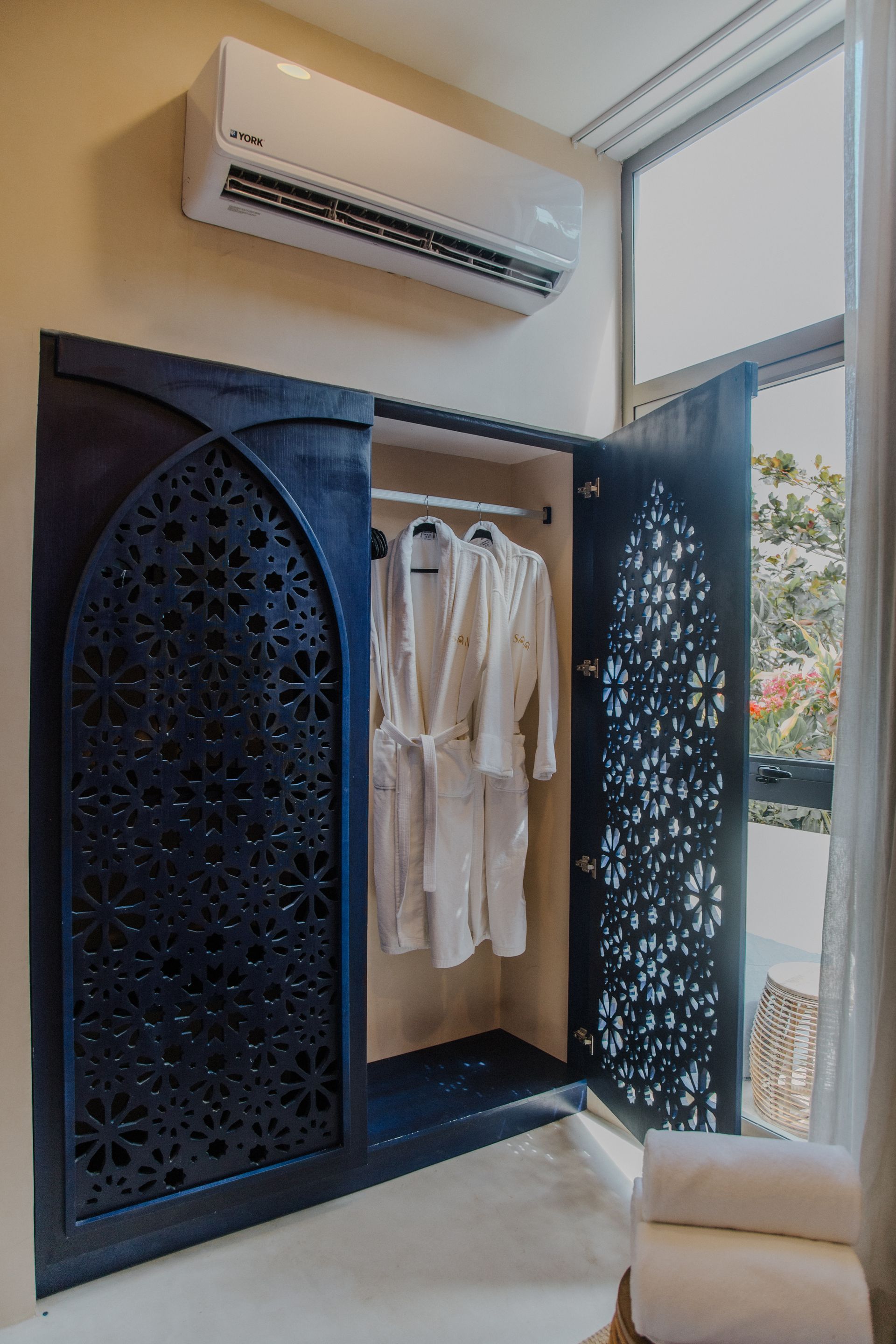 Un armario con puertas talladas de color azul oscuro y ornamentadas, que contiene dos batas de baño blancas, situado debajo de un aire acondicionado montado en la pared.