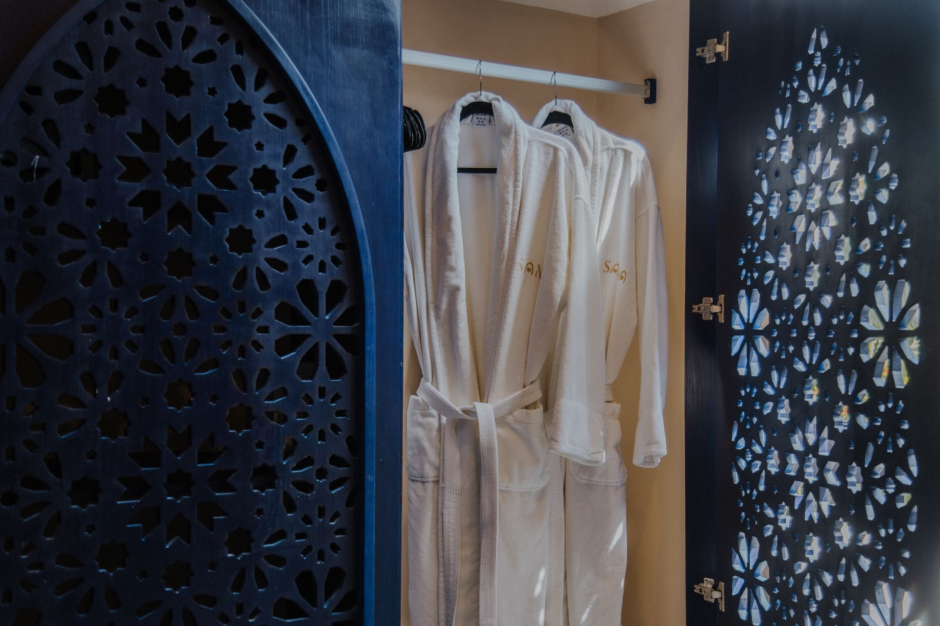 Dos batas blancas colgadas en un armario abierto con puertas de celosía geométrica de color azul oscuro y ornamentadas.