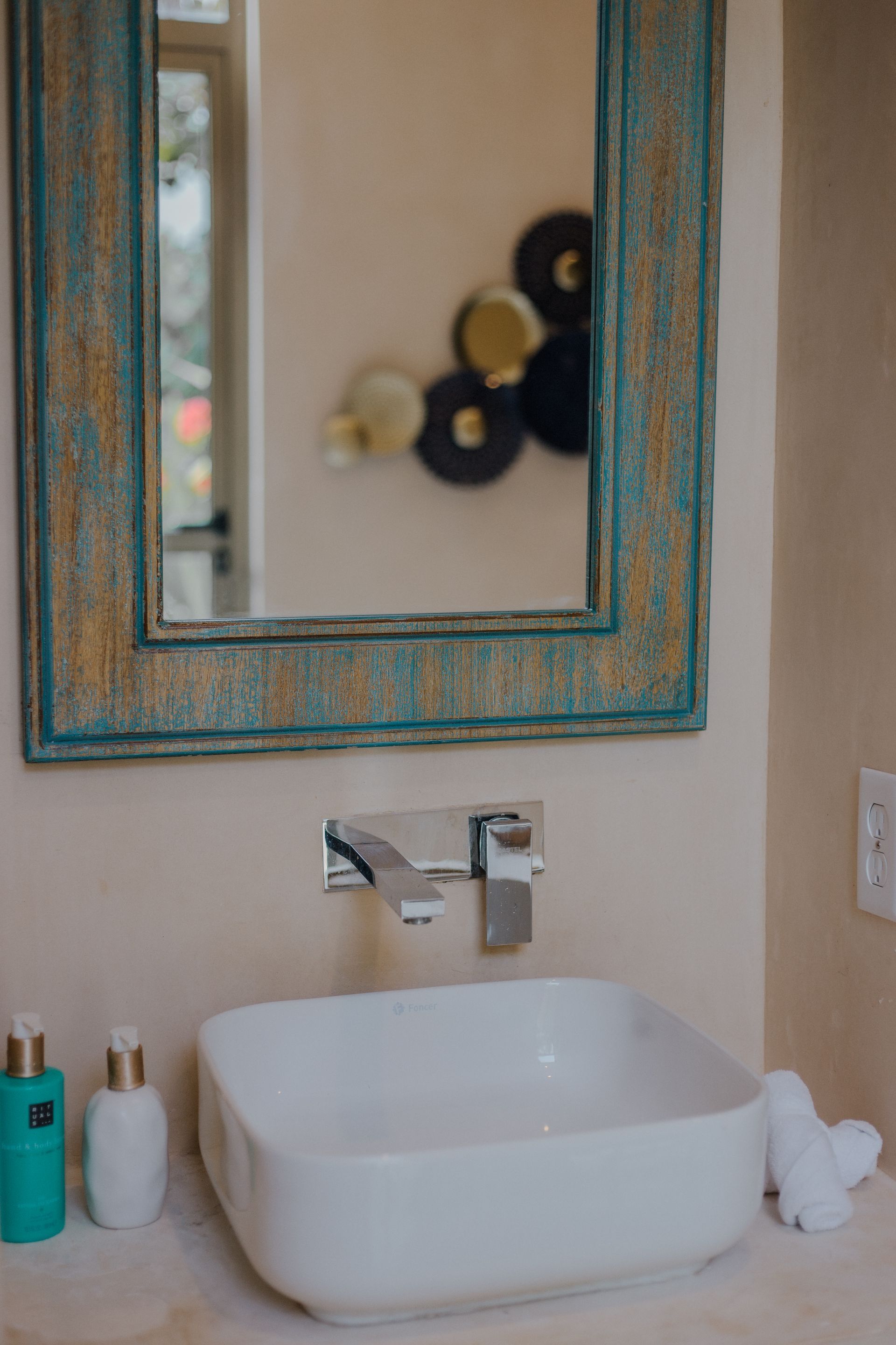 Un lavabo cuadrado blanco con un grifo cromado moderno, bajo un espejo rústico con marco turquesa y dorado.