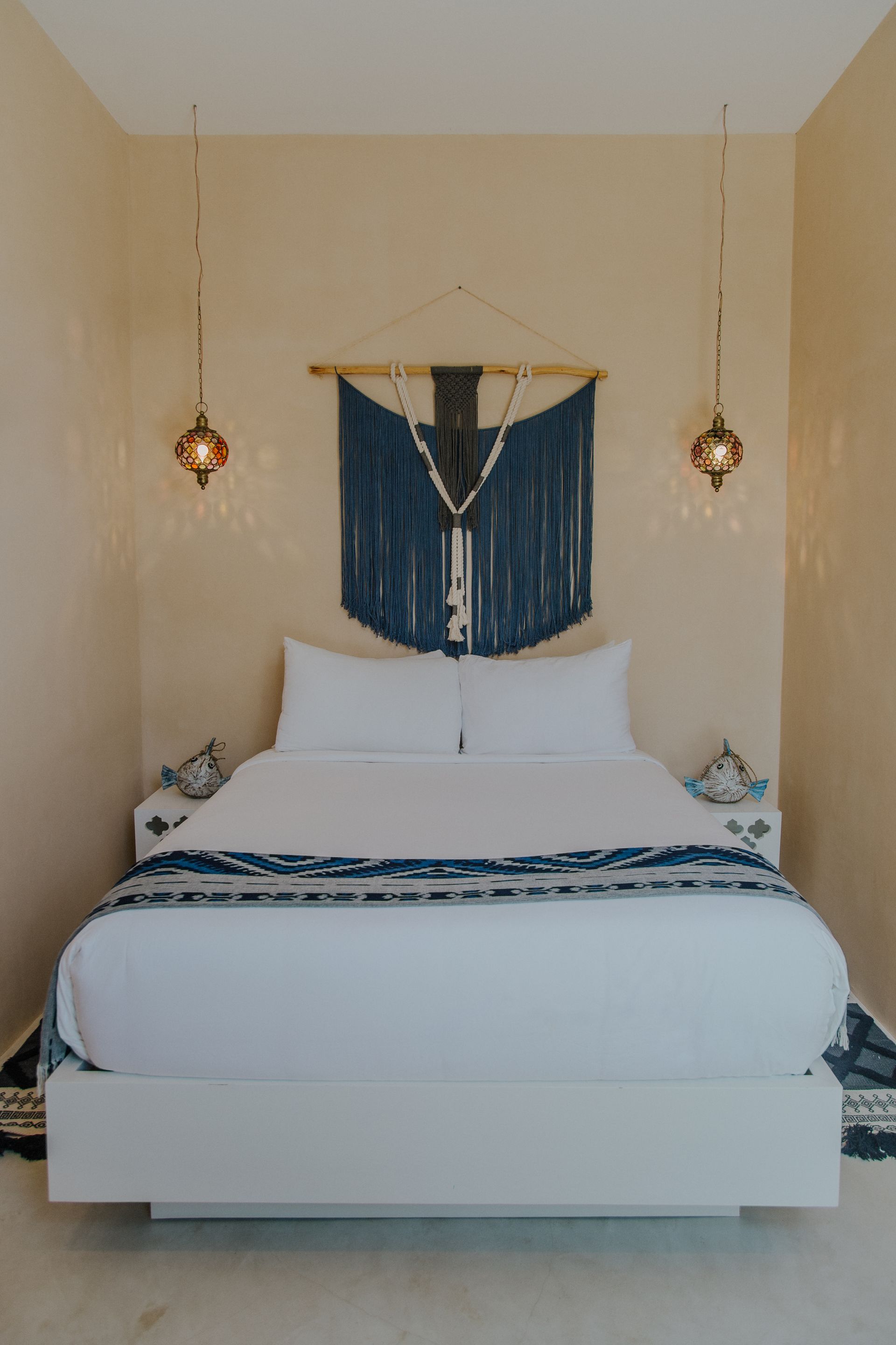 A bedroom features a white bed, a dark blue macramé wall hanging, and two hanging ornate gold pendant lights.