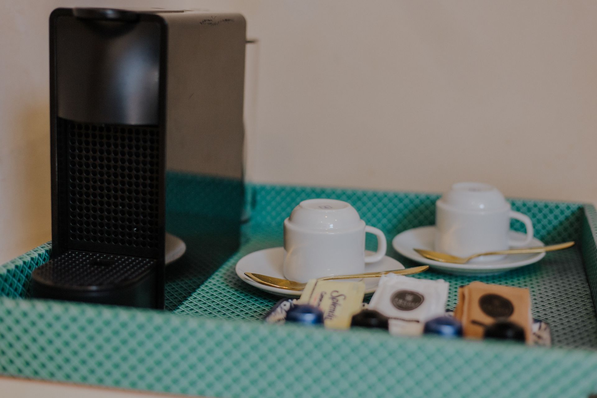 Una cafetera sobre una bandeja color turquesa con dos tazas blancas, platillos, cucharas doradas y cápsulas de café.