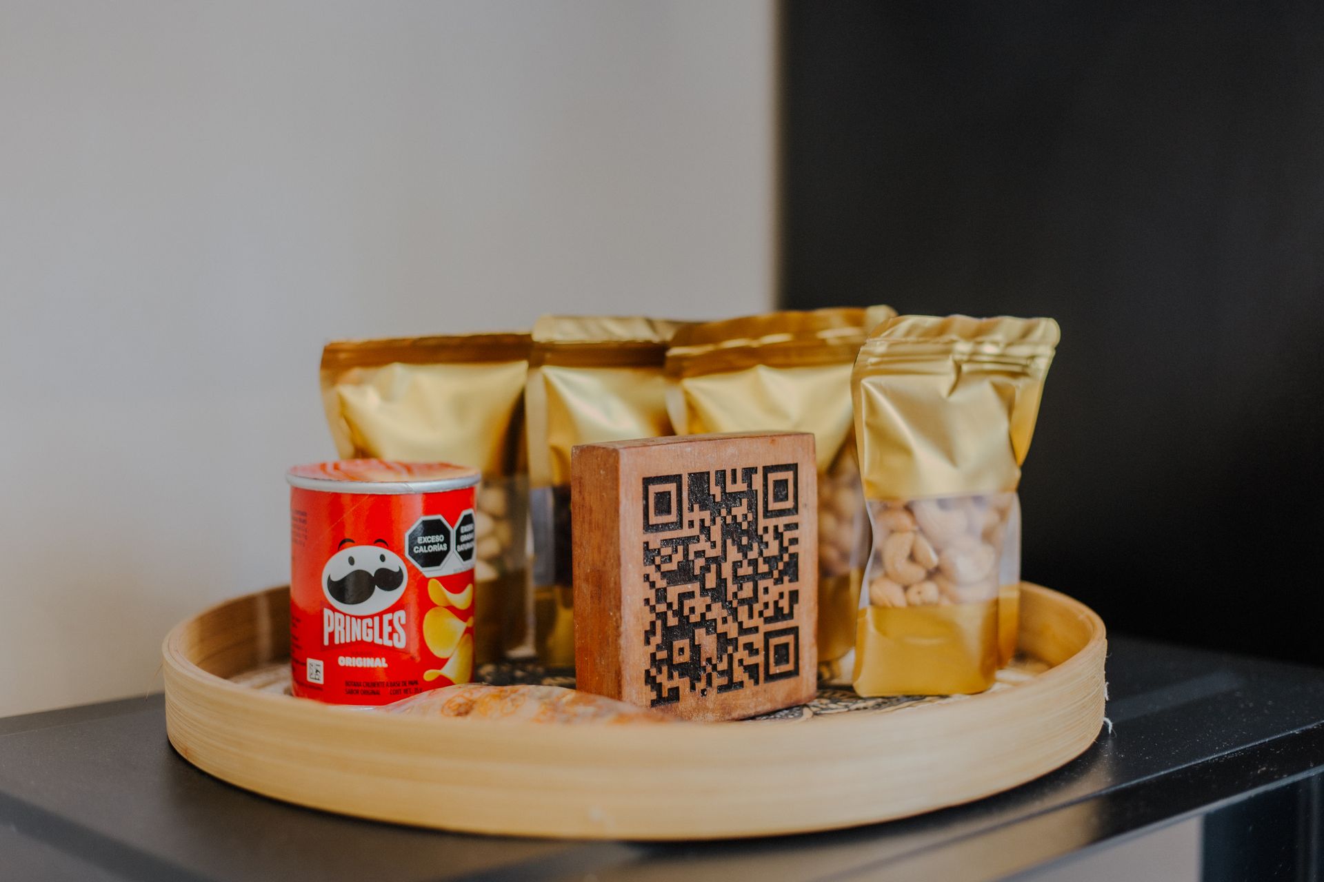 Una bandeja de bambú con una lata pequeña de Pringles, bolsas de aperitivos y un bloque cuadrado impreso con un código QR, sobre un mostrador.