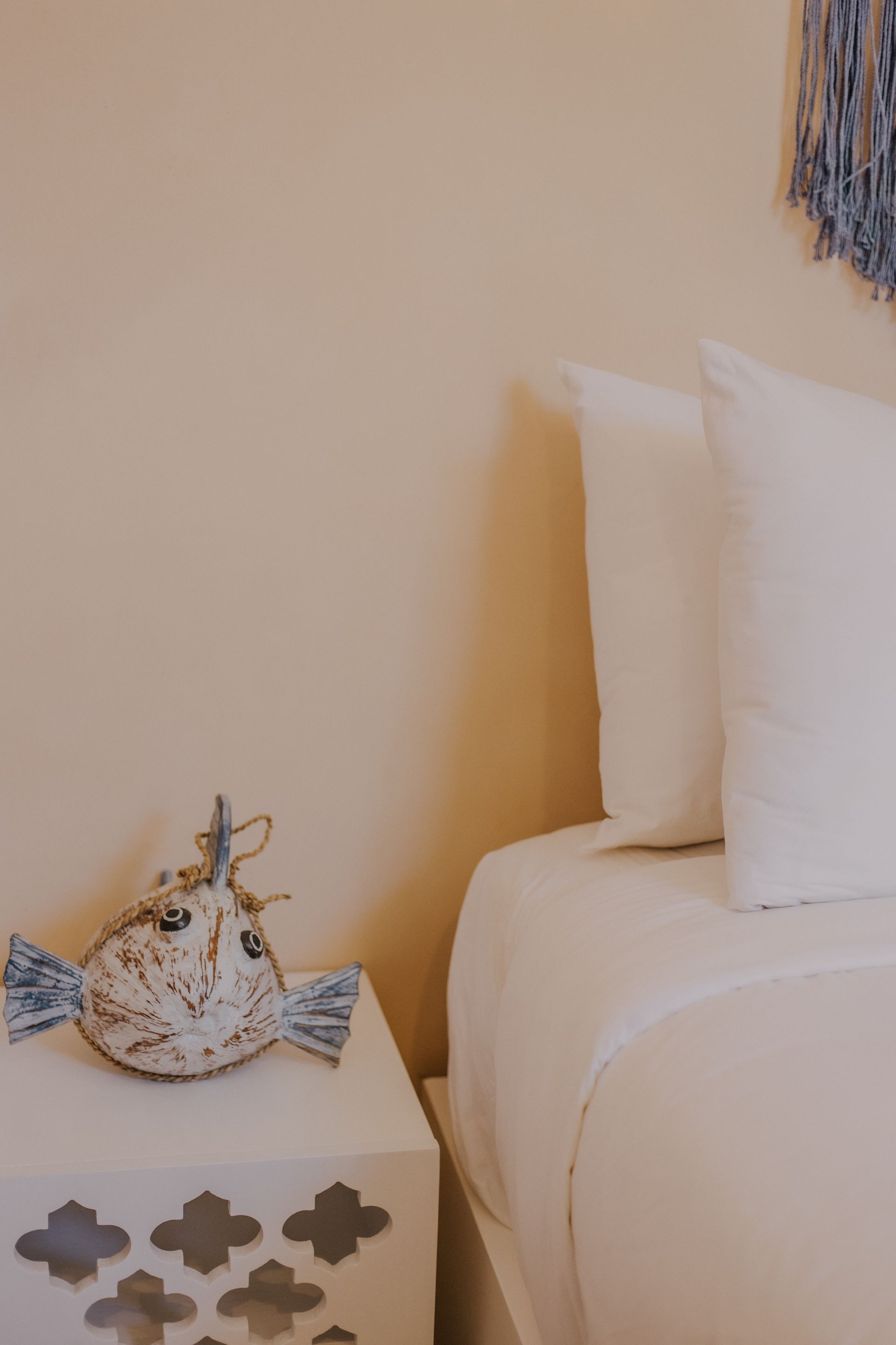 Un pez de peluche decorativo sobre una mesita de noche blanca estampada, junto a una cama con almohadas blancas y una pared color crema.