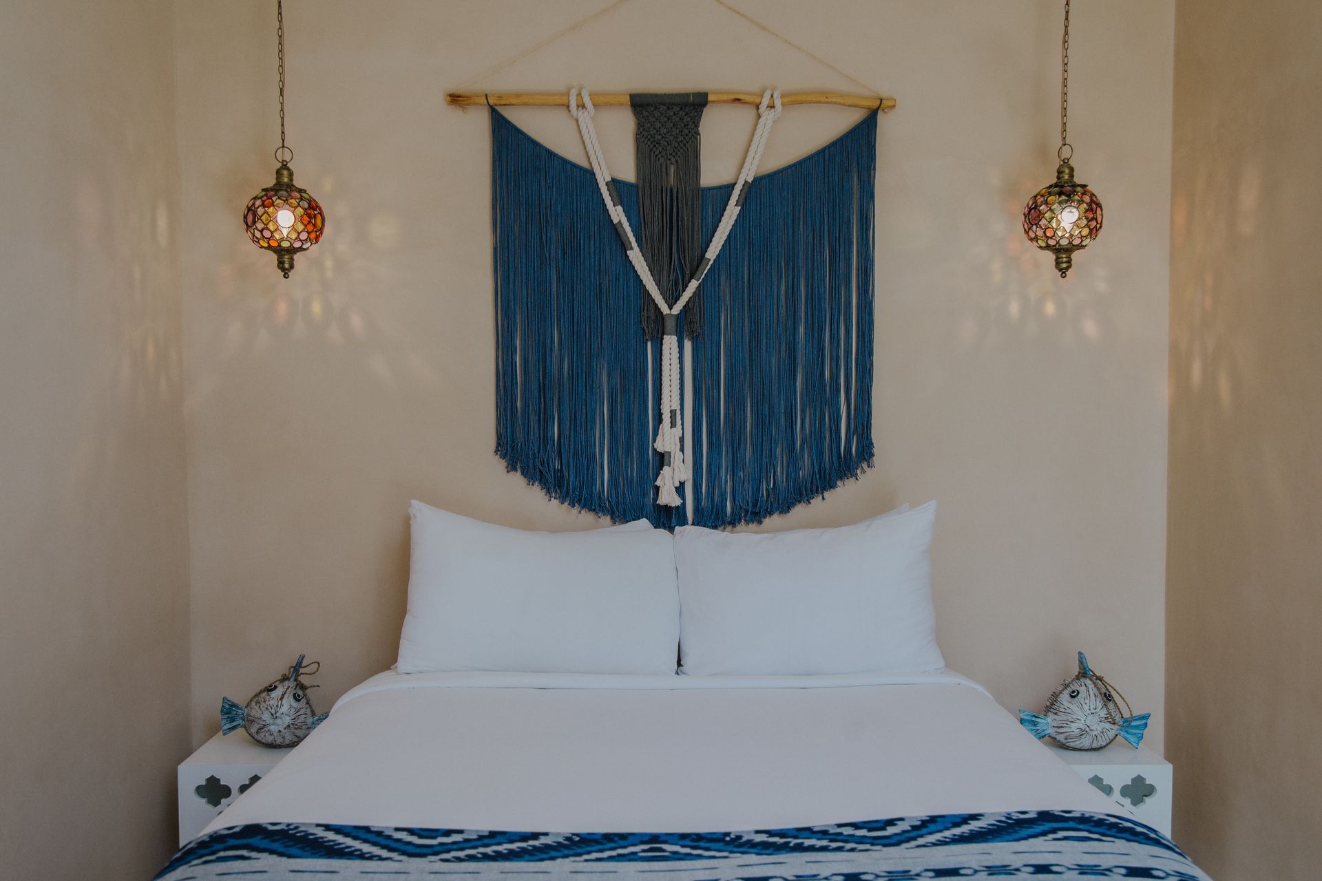 Una cama impecablemente hecha con almohadas blancas y una manta azul estampada, enmarcada por faroles metálicos colgantes y adornos de macramé azul.