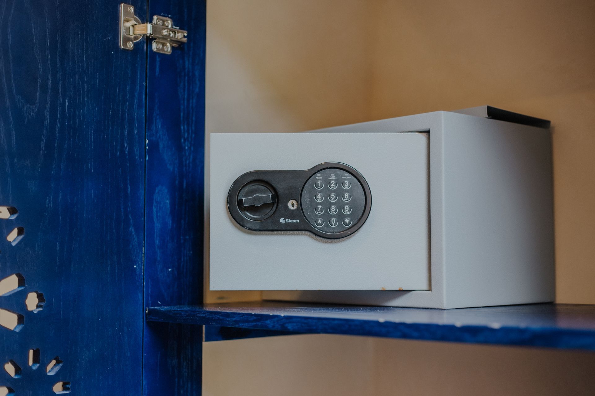 Una caja fuerte electrónica de color gris claro está abierta dentro de un armario azul con una puerta decorativa calada.