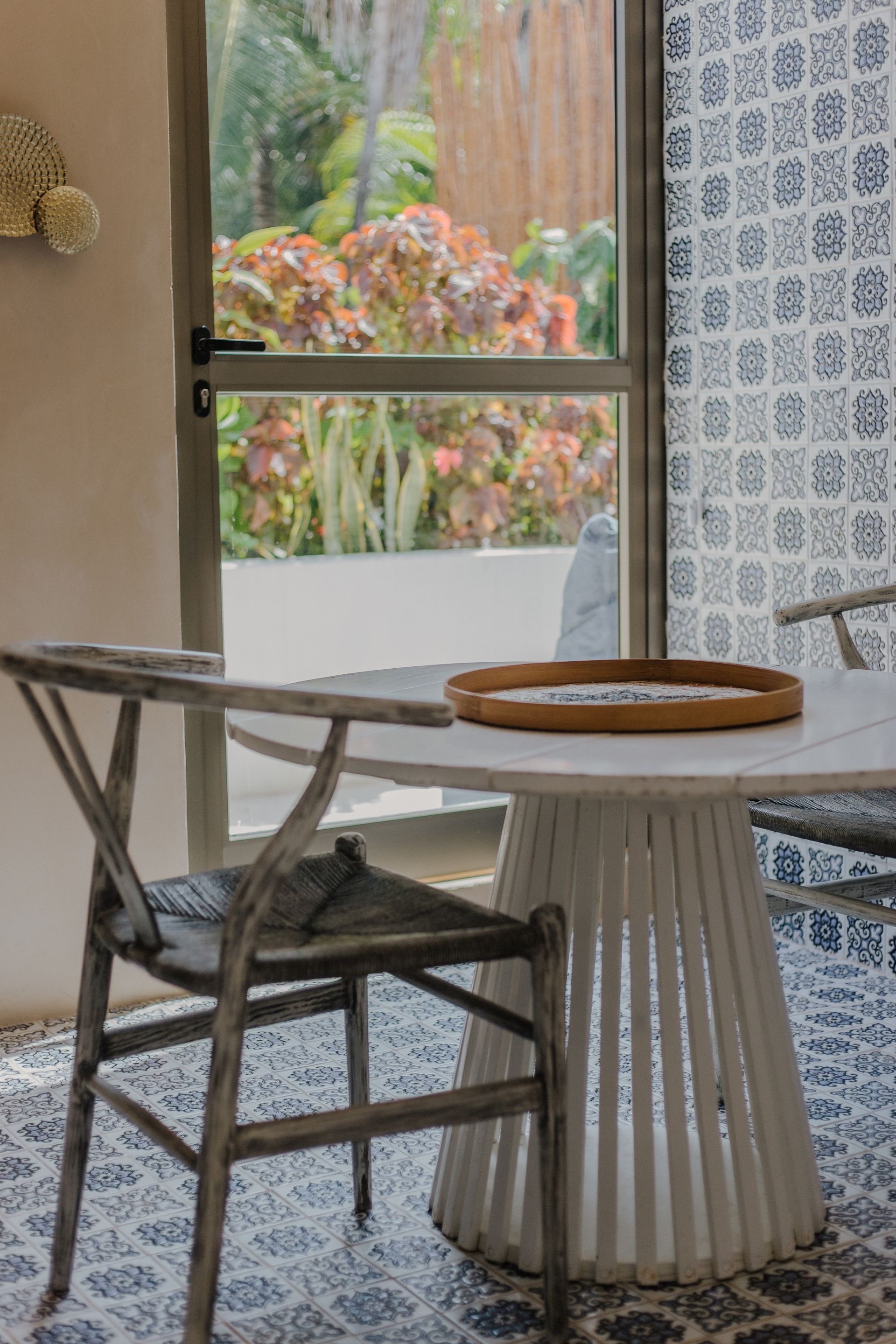 Una silla de madera desgastada se encuentra junto a una mesa blanca con pedestal sobre un suelo de baldosas con estampado azul, frente a una puerta de cristal.