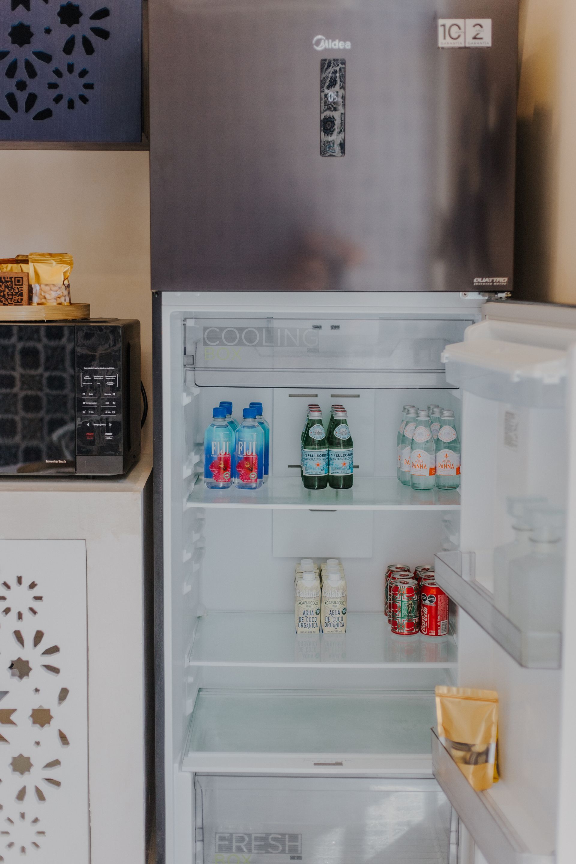 En una cocina, cerca de un microondas, hay un frigorífico Midea abierto, lleno de botellas de agua, refrescos y aperitivos.