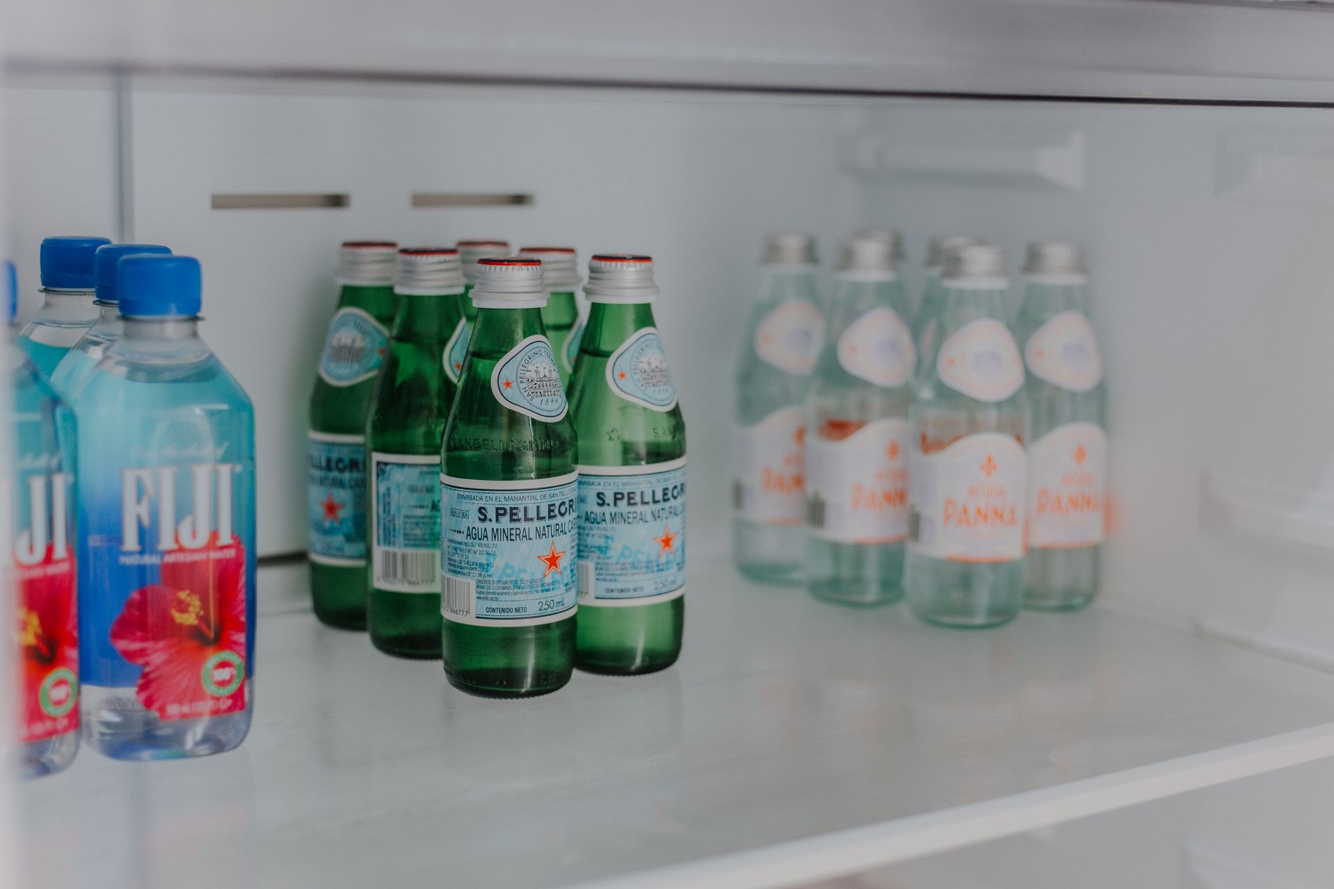 Dentro de un estante blanco del refrigerador hay varias botellas de vidrio de agua con gas San Pellegrino y dos botellas de agua Fiji.