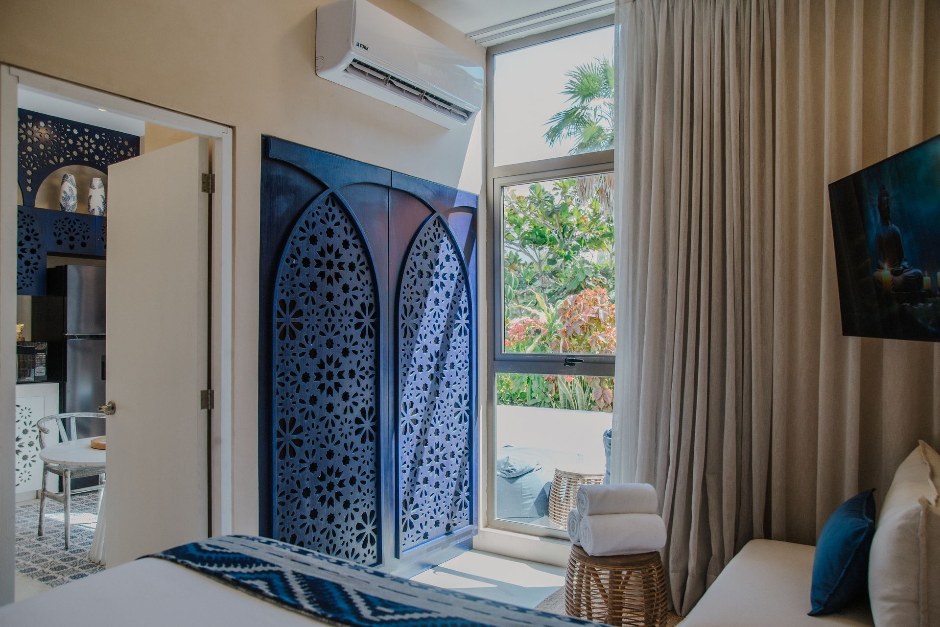 Una habitación de hotel con un armario azul ornamentado, una gran ventana con cortinas que llegan hasta el suelo y vistas a la vegetación exterior.