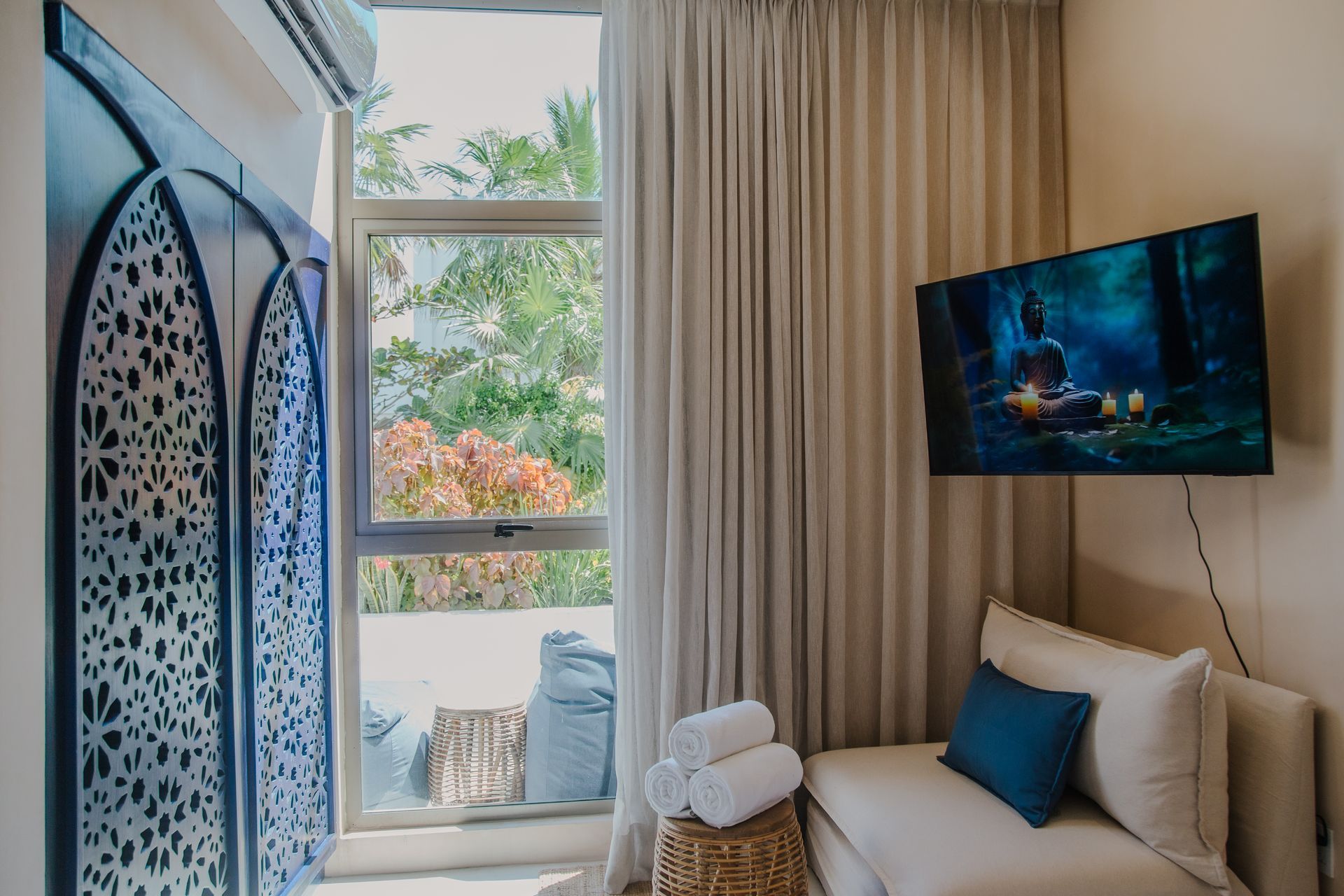 Una habitación con un biombo azul estampado, una ventana con vistas a la vegetación y un pequeño sofá con un televisor montado encima.