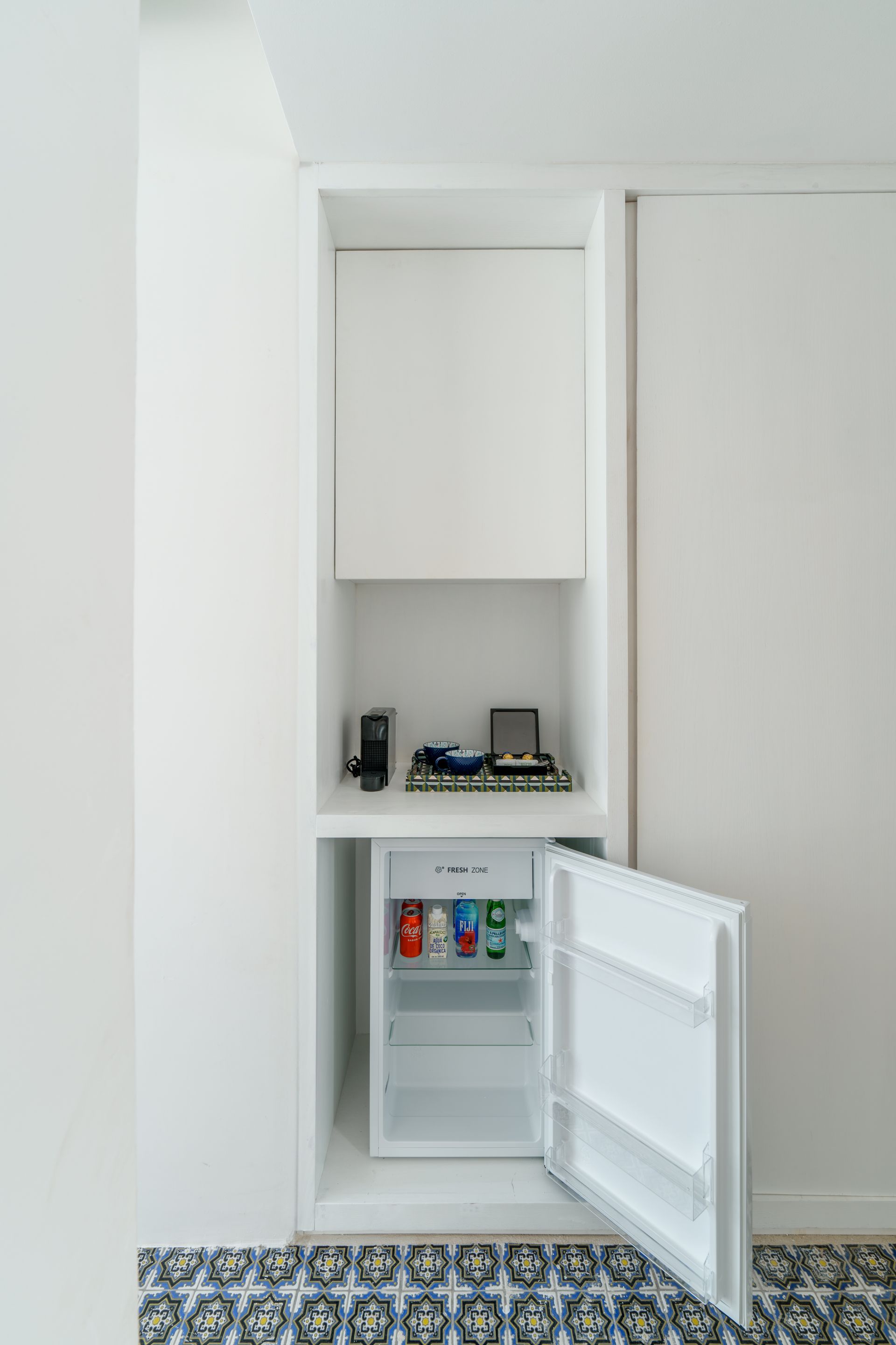 Mueble blanco empotrado con mininevera abierta, repleta de bebidas, y estante para aparatos electrónicos.