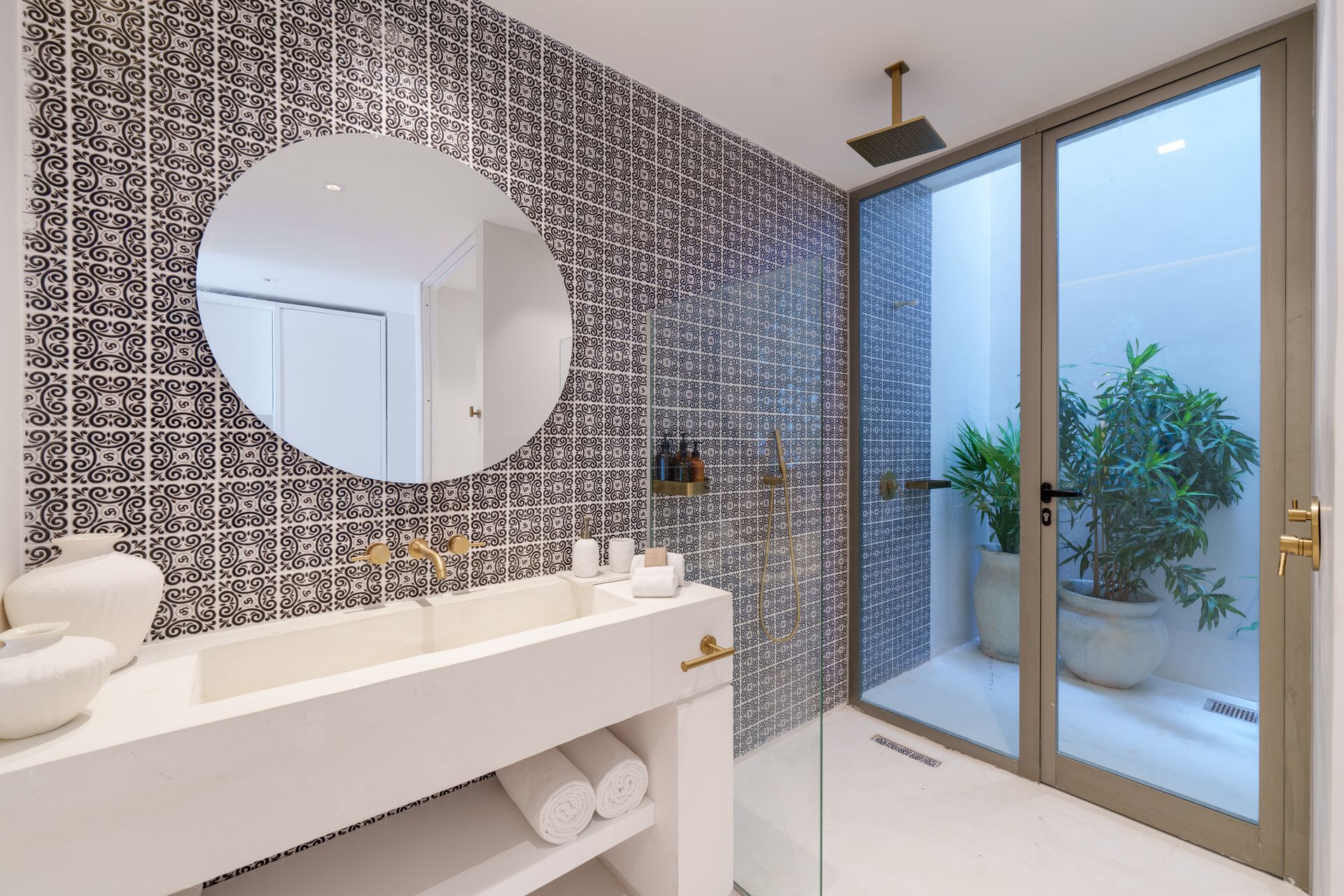 Baño con azulejos estampados en azul y blanco, lavabo blanco, espejo redondo y una puerta de vidrio que conduce a un patio con una planta en maceta.