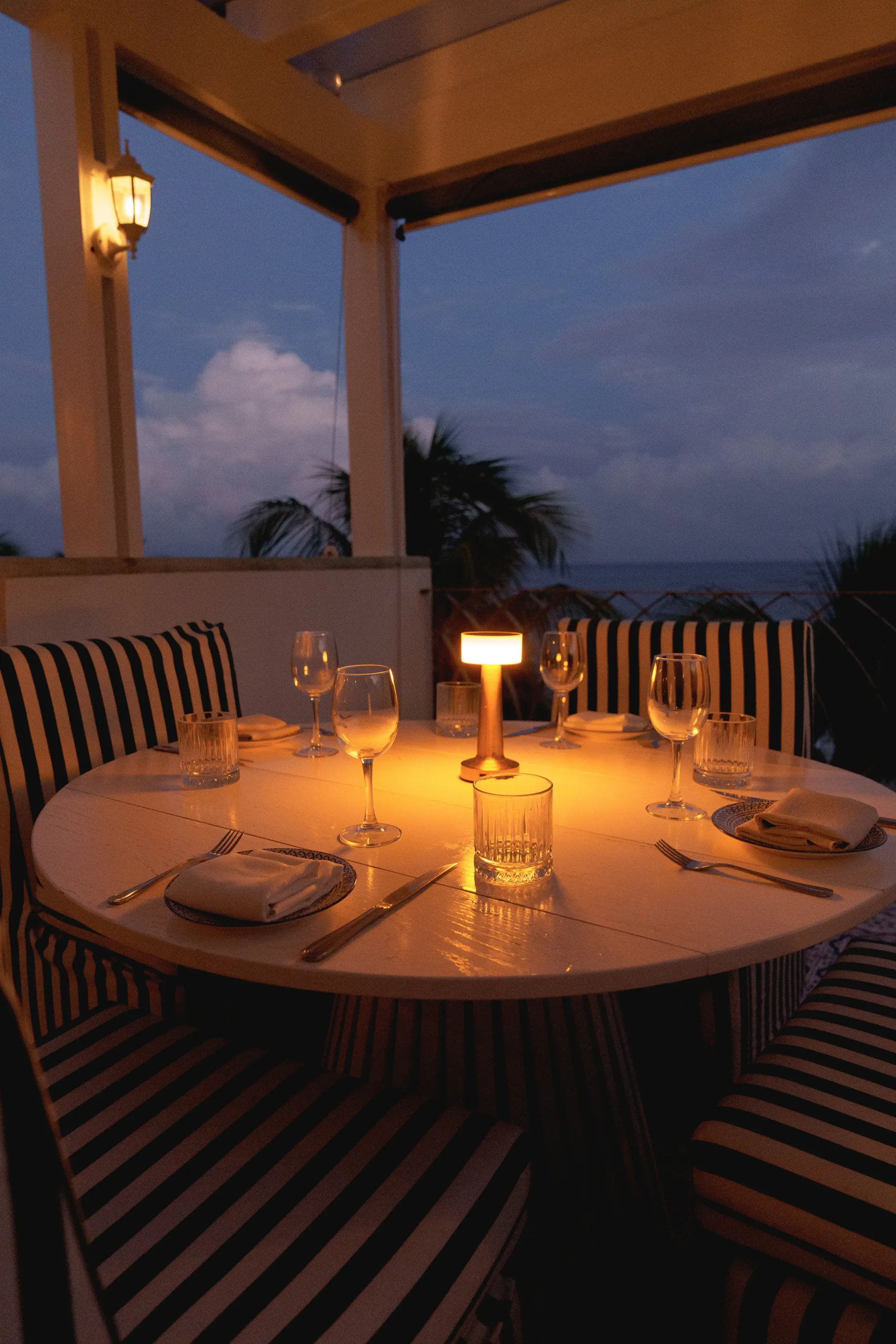 Una mesa de comedor circular para cuatro personas en una terraza al aire libre al atardecer, iluminada por una cálida lámpara de mesa central.