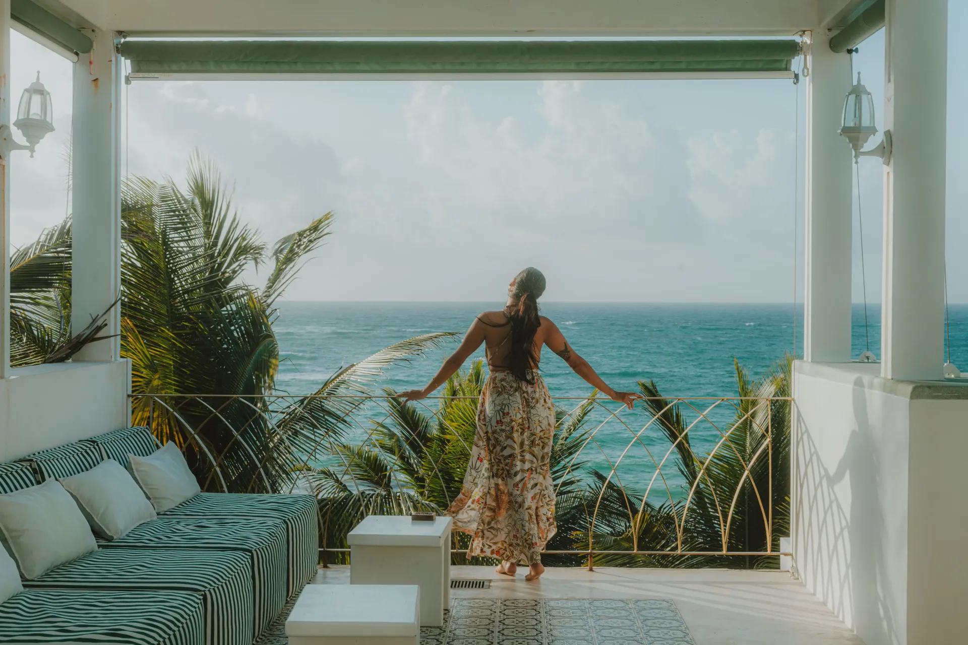 Una persona se encuentra en un balcón con vistas al océano, con los brazos extendidos, luciendo un vestido largo estampado.