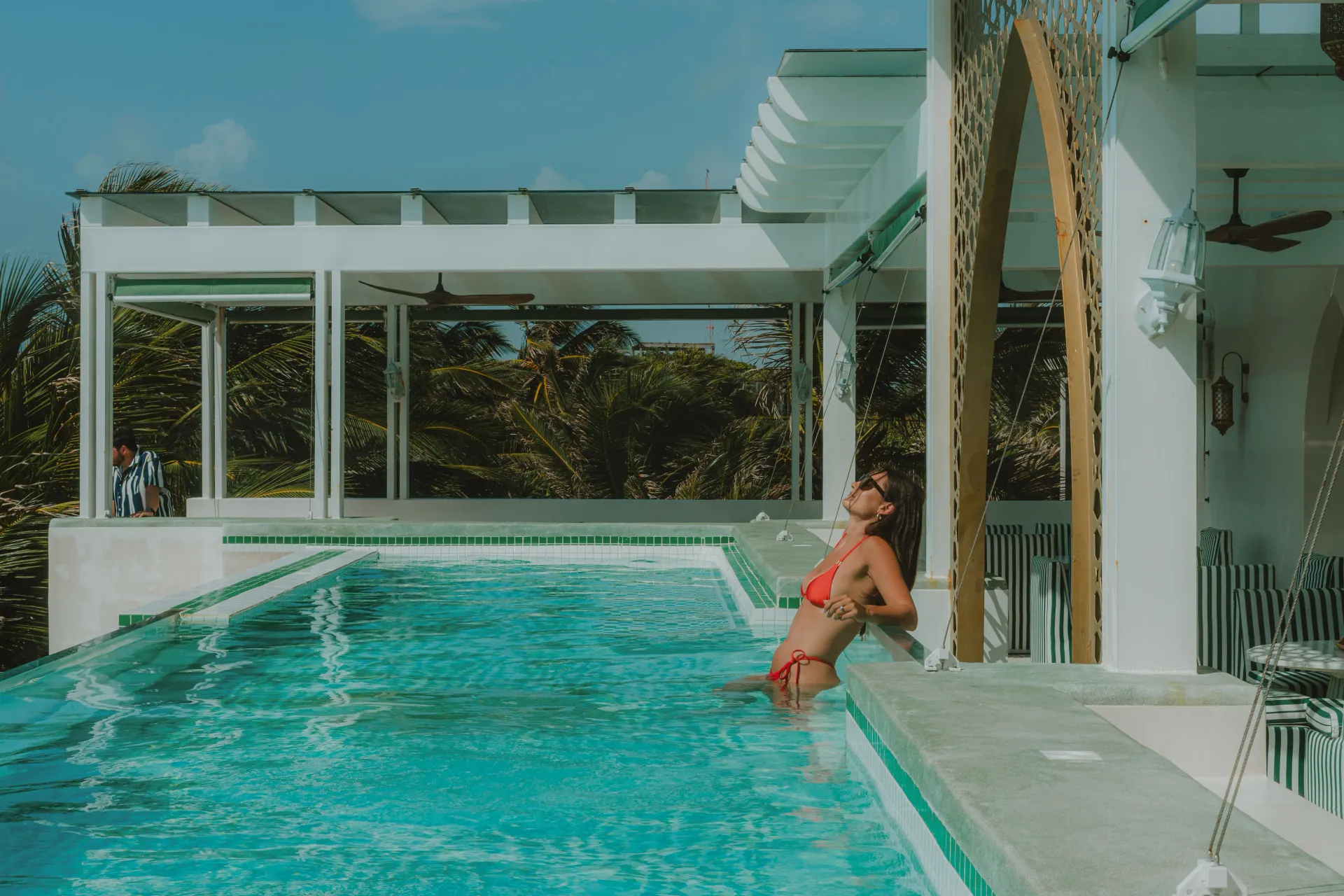 Una persona en bikini rojo se apoya en el borde de una piscina infinita de color turquesa brillante en una villa soleada con columnas blancas.