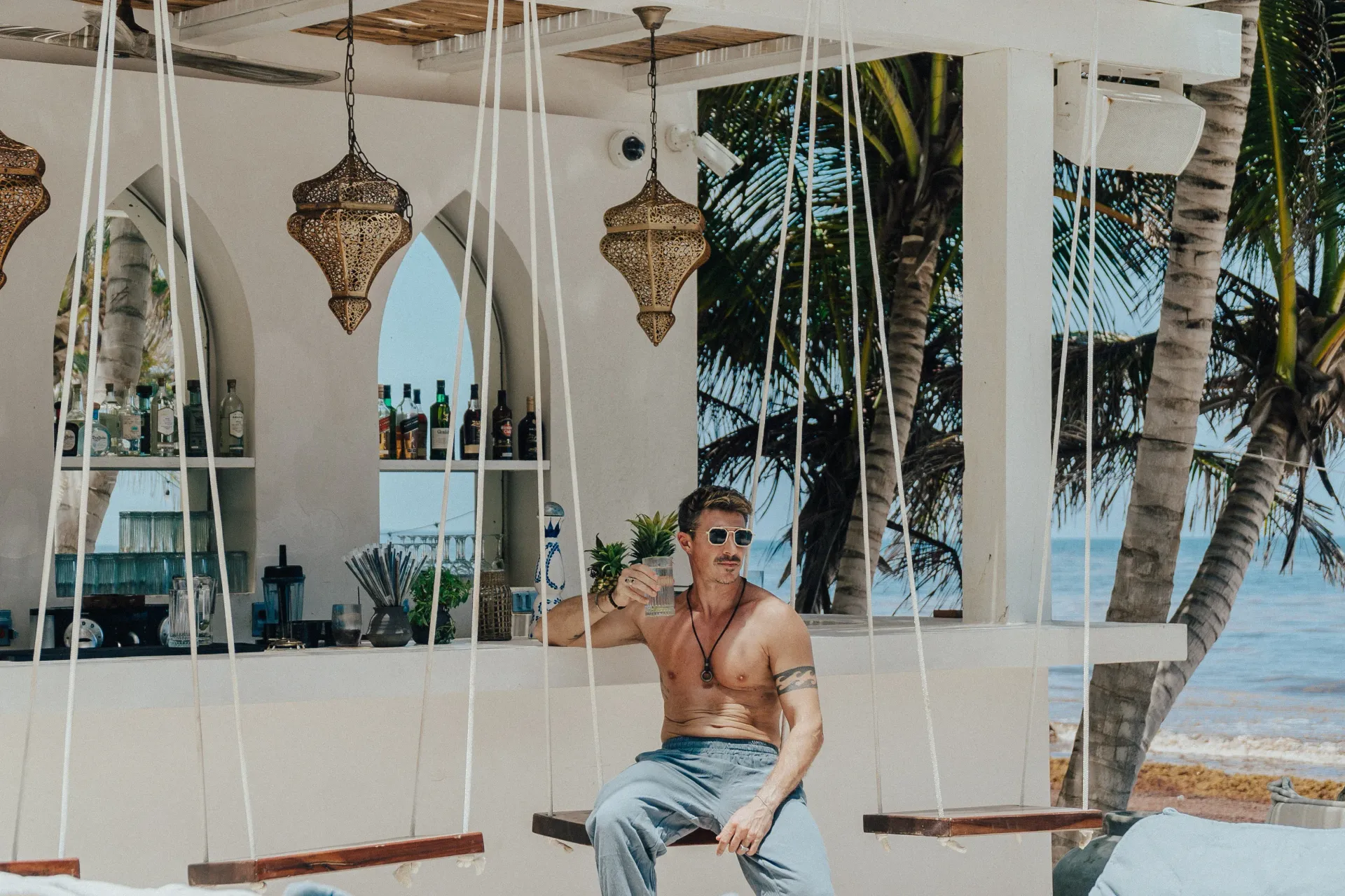 A bare-chested person sits on a swing at an open-air beach bar with hanging lamps and bottles on shelves behind them.