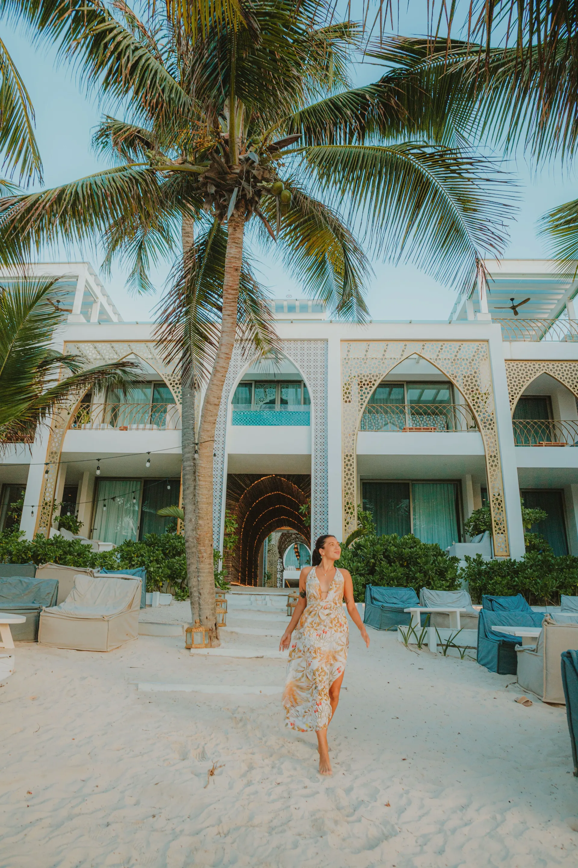 A person in a patterned dress walking on a white sandy beach toward a white resort building with arched walkways.