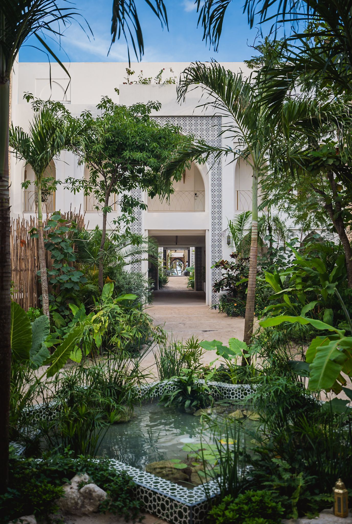 Patio con exuberante vegetación, un estanque reflectante y un edificio blanco con arcos.