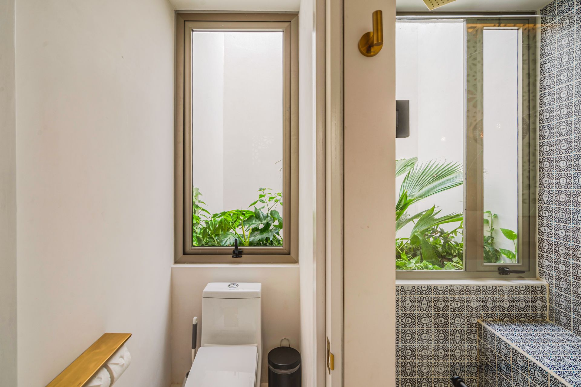 Baño con inodoro, ventanas que dan a un pequeño jardín y azulejos estampados.