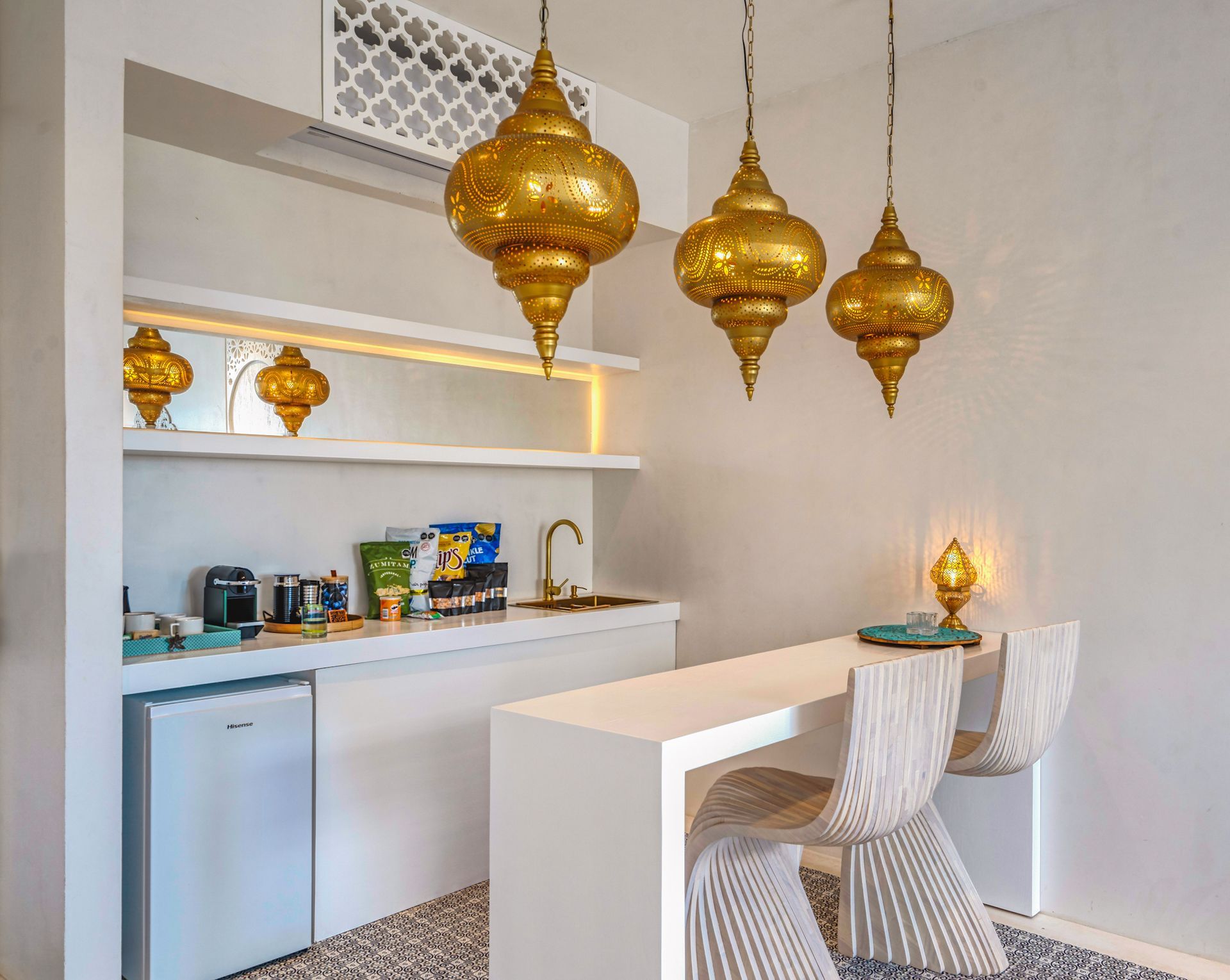 Un área de bar de color blanco brillante con luces colgantes doradas, sillas blancas y un refrigerador pequeño.