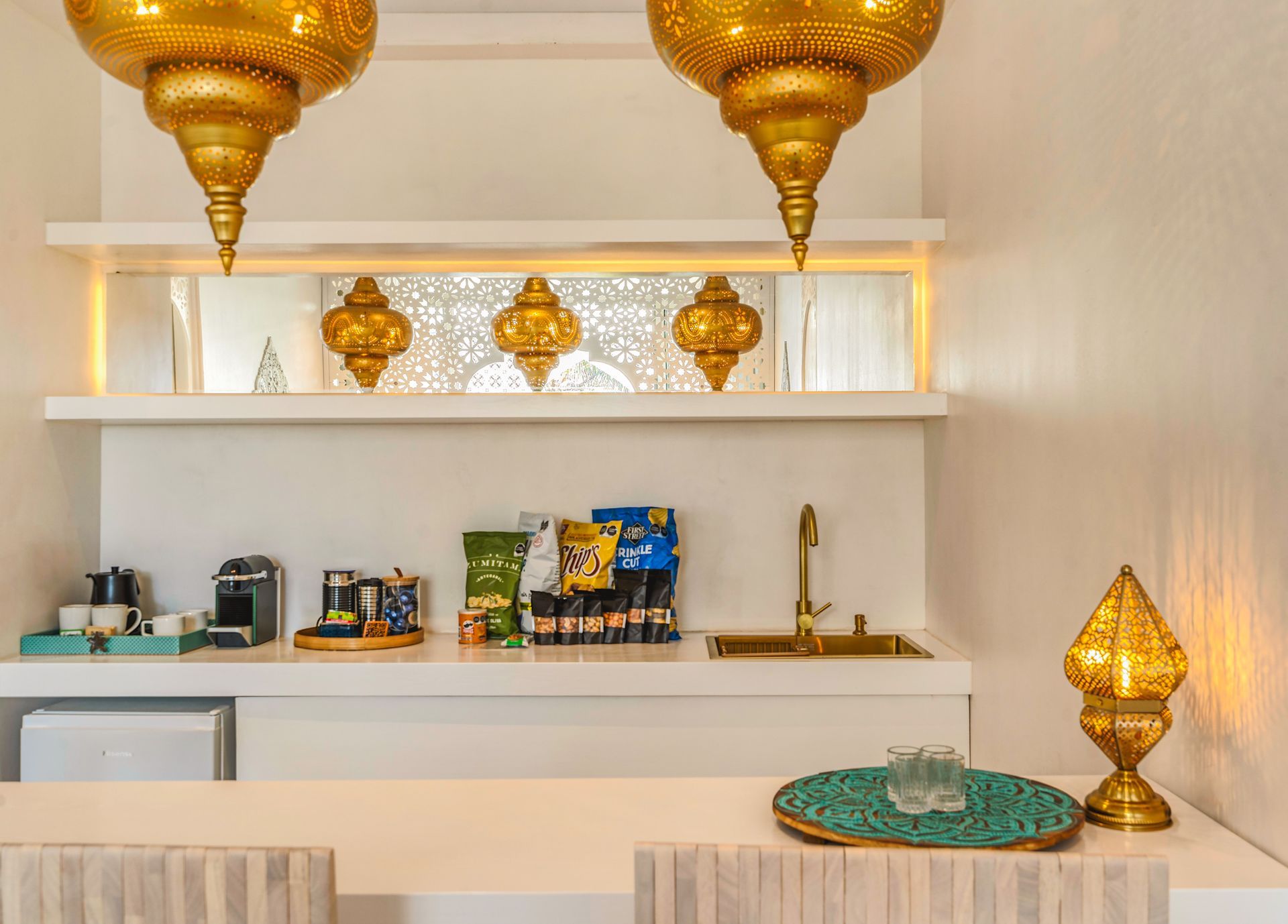 Área de bar con lámparas doradas ornamentadas, estanterías blancas con bocadillos, fregadero y bandeja verde azulado.
