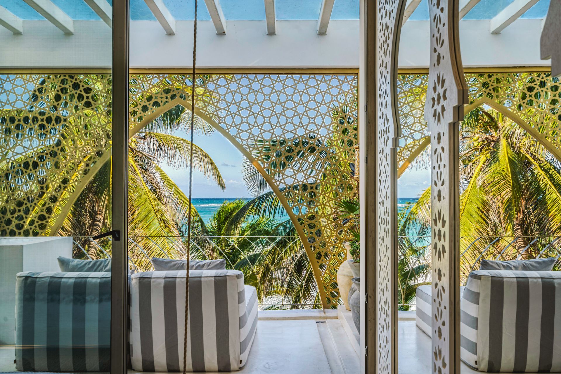 Balcony with striped seating, ocean view through ornate screen, palm trees.