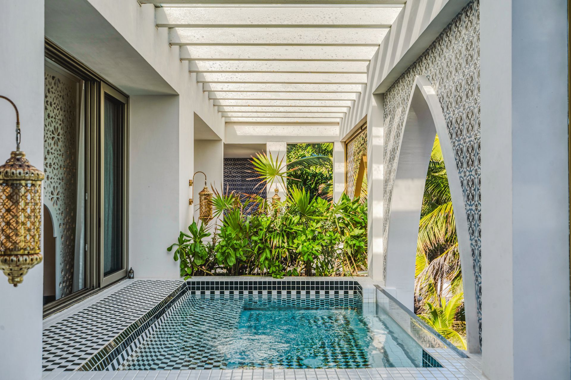 Una pequeña área de piscina con azulejos, una pérgola blanca, detalles de azulejos estampados y una exuberante vegetación.